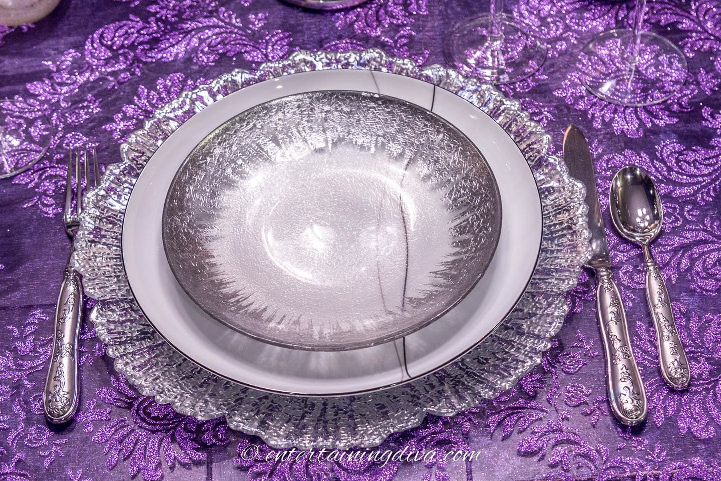 Place setting with a silver salad plate, black and white dinner plate and silver charger on a purple lace tablecloth