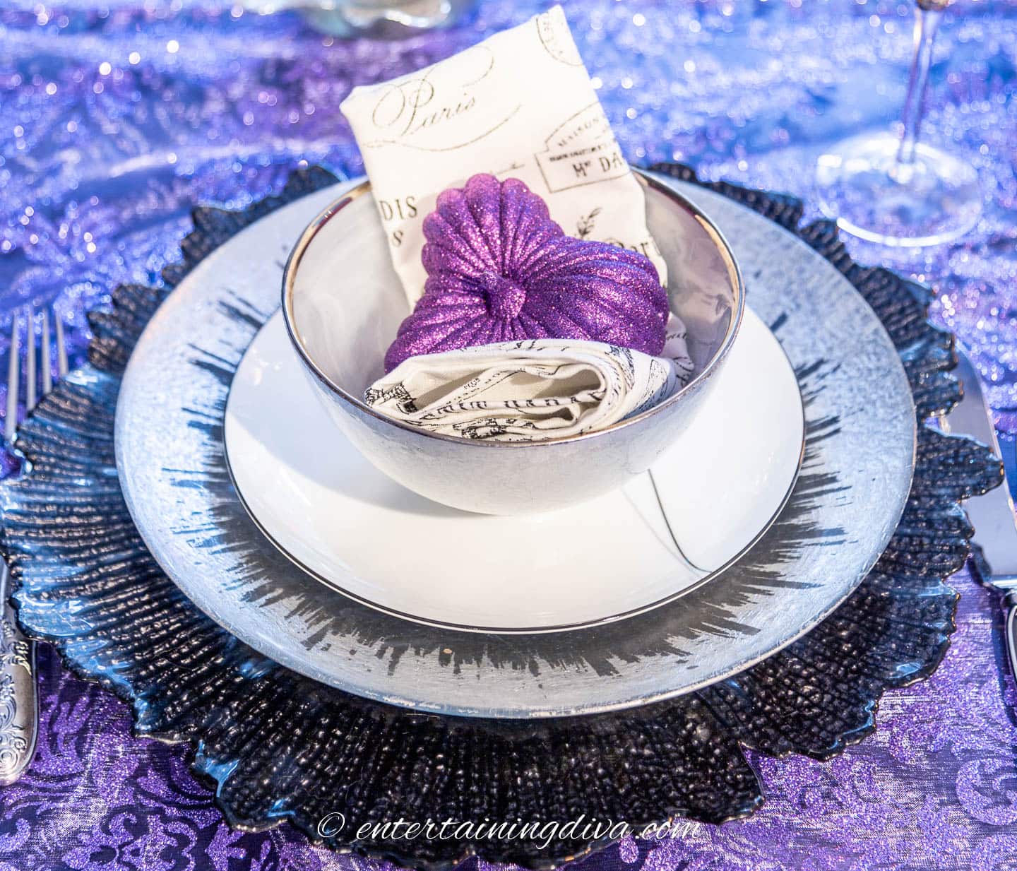 Elegant Halloween place setting with a faux purple pumpkin, black and white napkins, silver and white plates on a black charger
