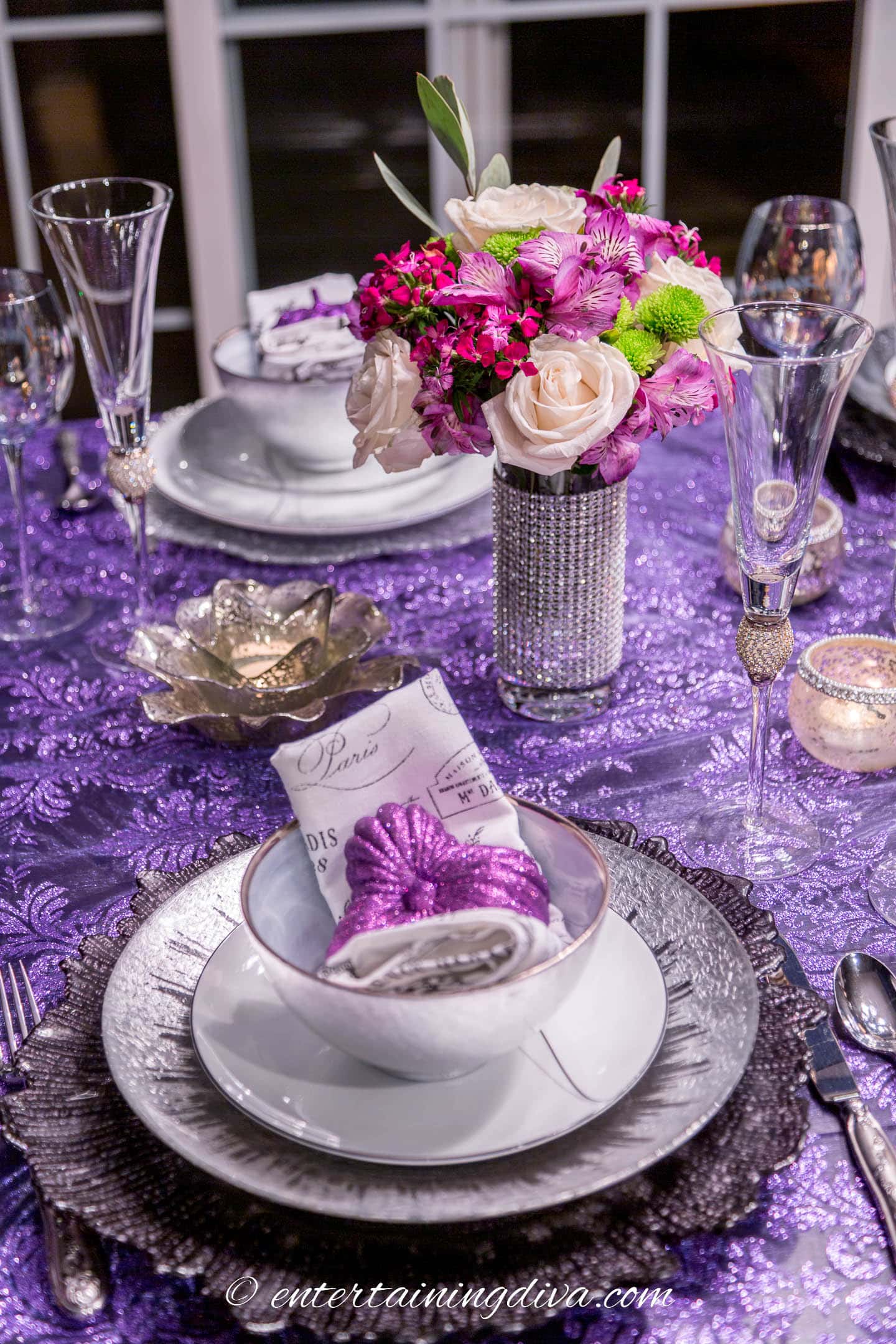 Black and silver place setting with a purple pumpkin on a purple lace tablecloth in front of a small flower arrangement