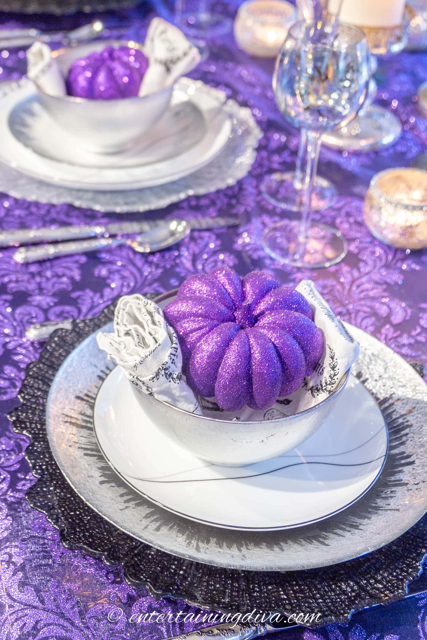 Place settings with small faux purple pumpkins in a bowl on top of a black and white napkin, black and white salad plate, silver dinner plate and black charger