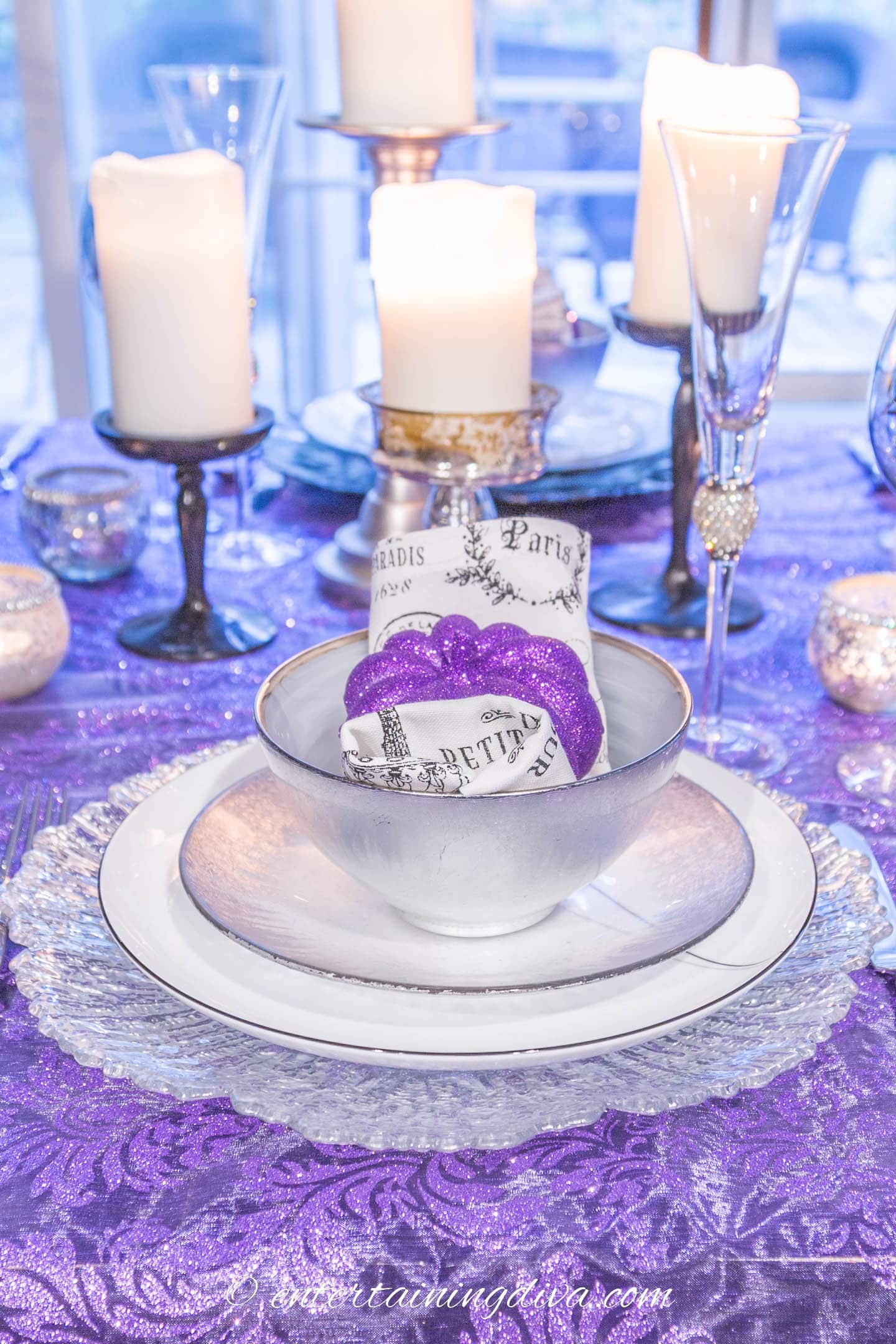 Place setting with silver and white dishes, black and white napkin and small purple pumpkin in front of some white candles