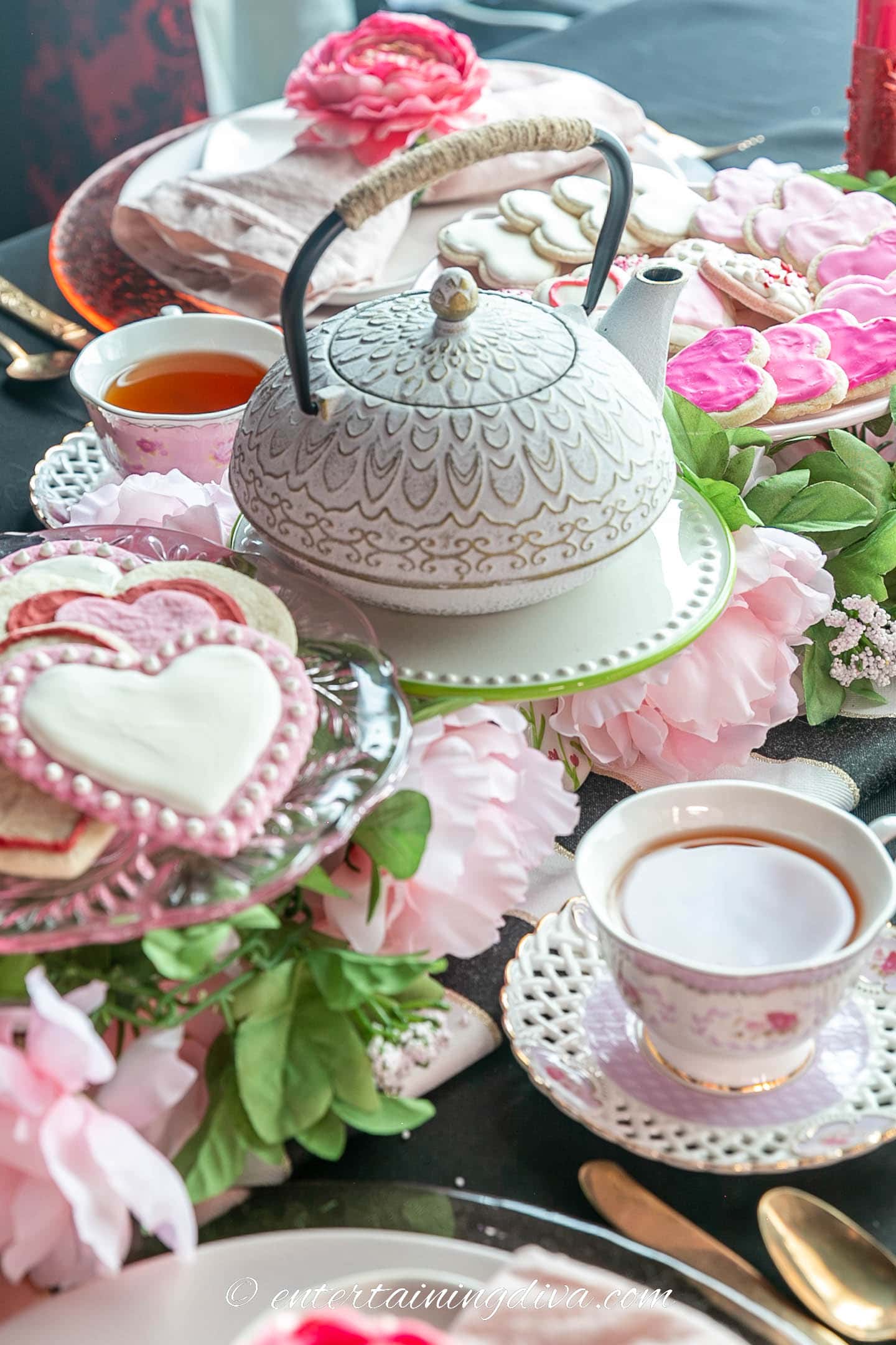 A teapot and heart cookies on cake stands above pink faux flowers as part of a Valentine's Day tea party centerpiece