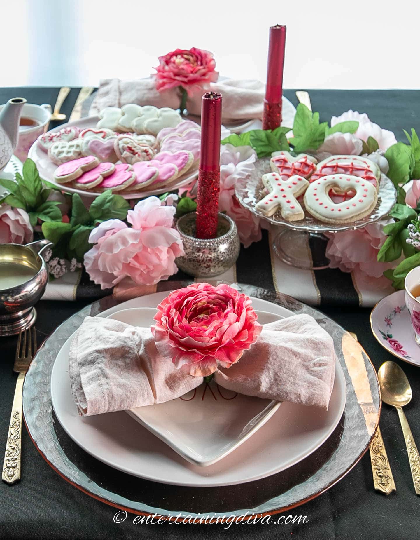 A Valentine place setting with a glass charger, light pink dinner plate, heart-shaped appetizer plate, linen napkin with flower napkin ring, gold cutlery and candles in the background