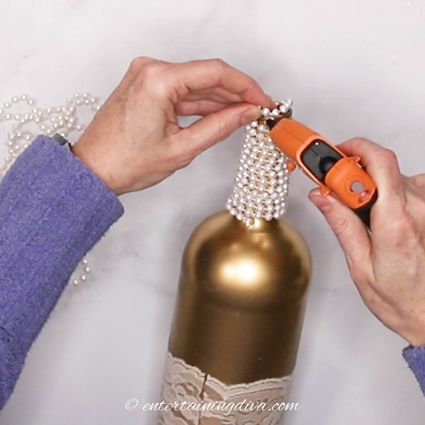 The end of a string of pearls being attached to the top of a wine bottle with a glue gun