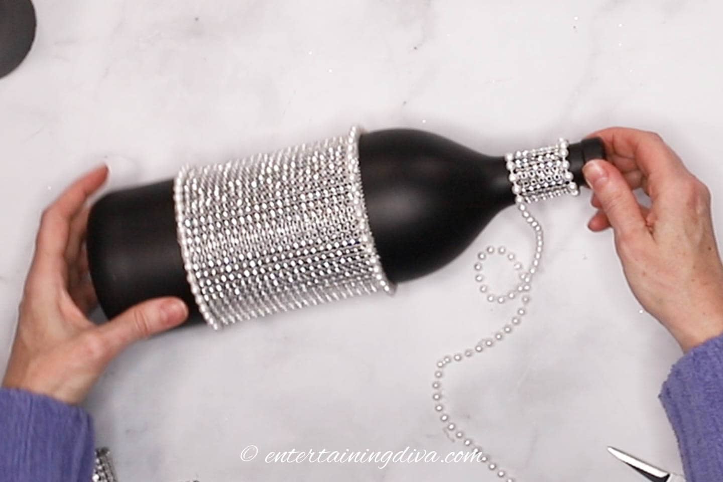 Faux pearls being glued to the bottom edge of silver ribbon wrapped around the neck of a black wine bottle