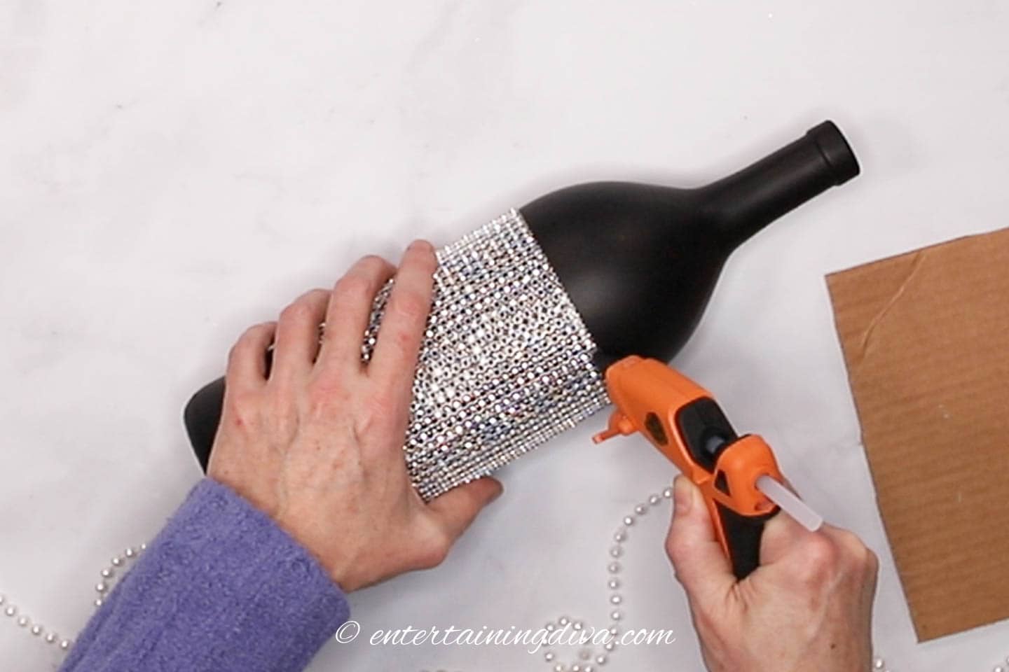 glue gun putting a line of glue on the edge of a rhinestone ribbon wrapped around a black wine bottle