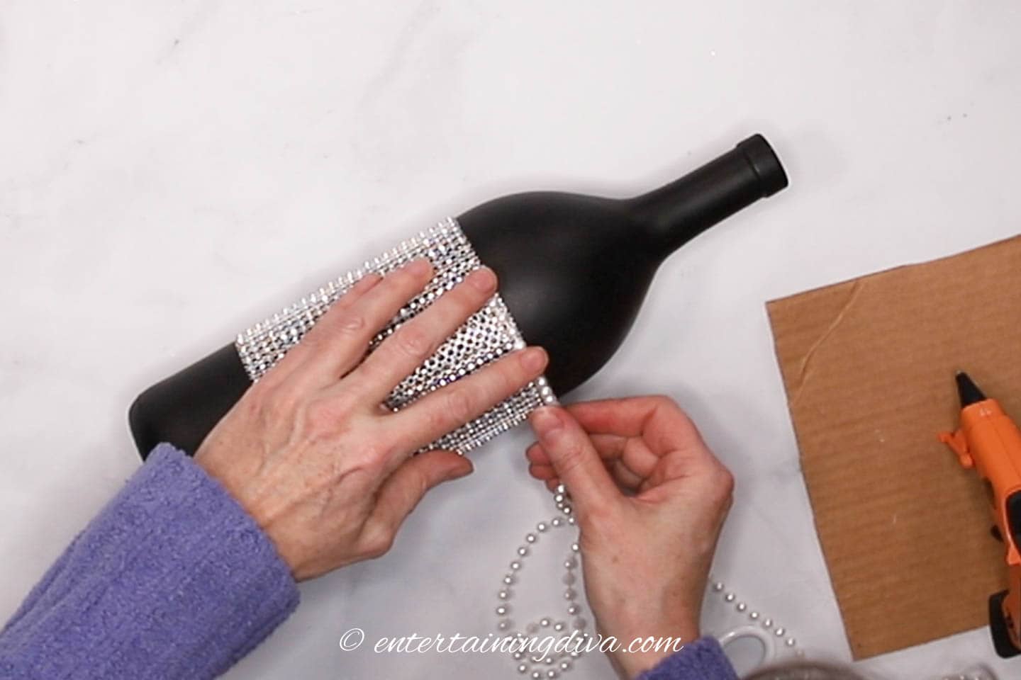 Faux pearls being stuck onto the top edge of silver ribbon that is wrapped around a wine bottle