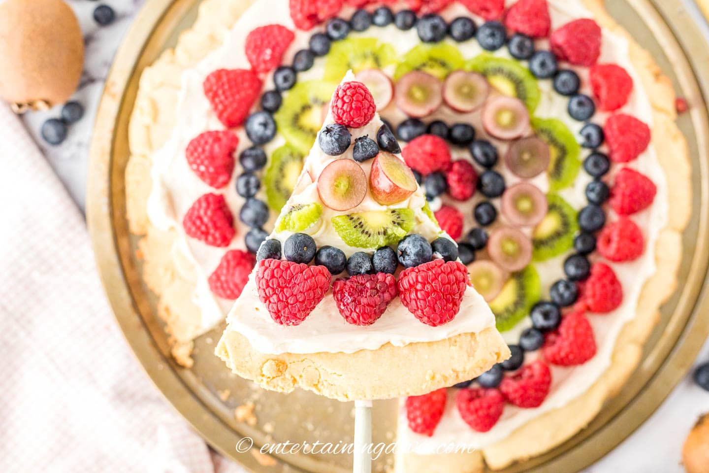 A slice of fruit pizza topped with raspberries, blueberries, kiwi and grapes being held above the rest of the pizza