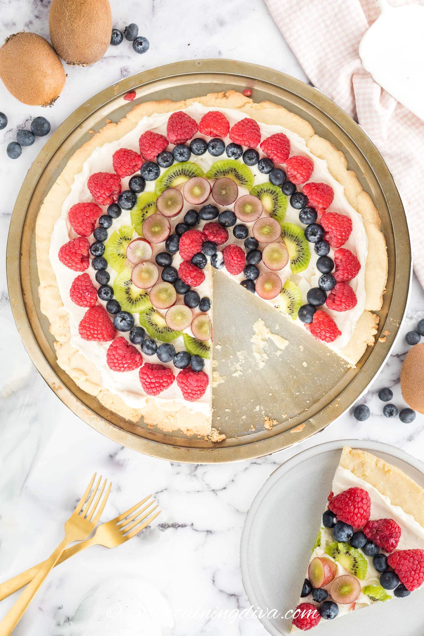 A slice of fruit pizza topped with raspberries, blueberries, kiwi and grapes being held above the rest of the pizza