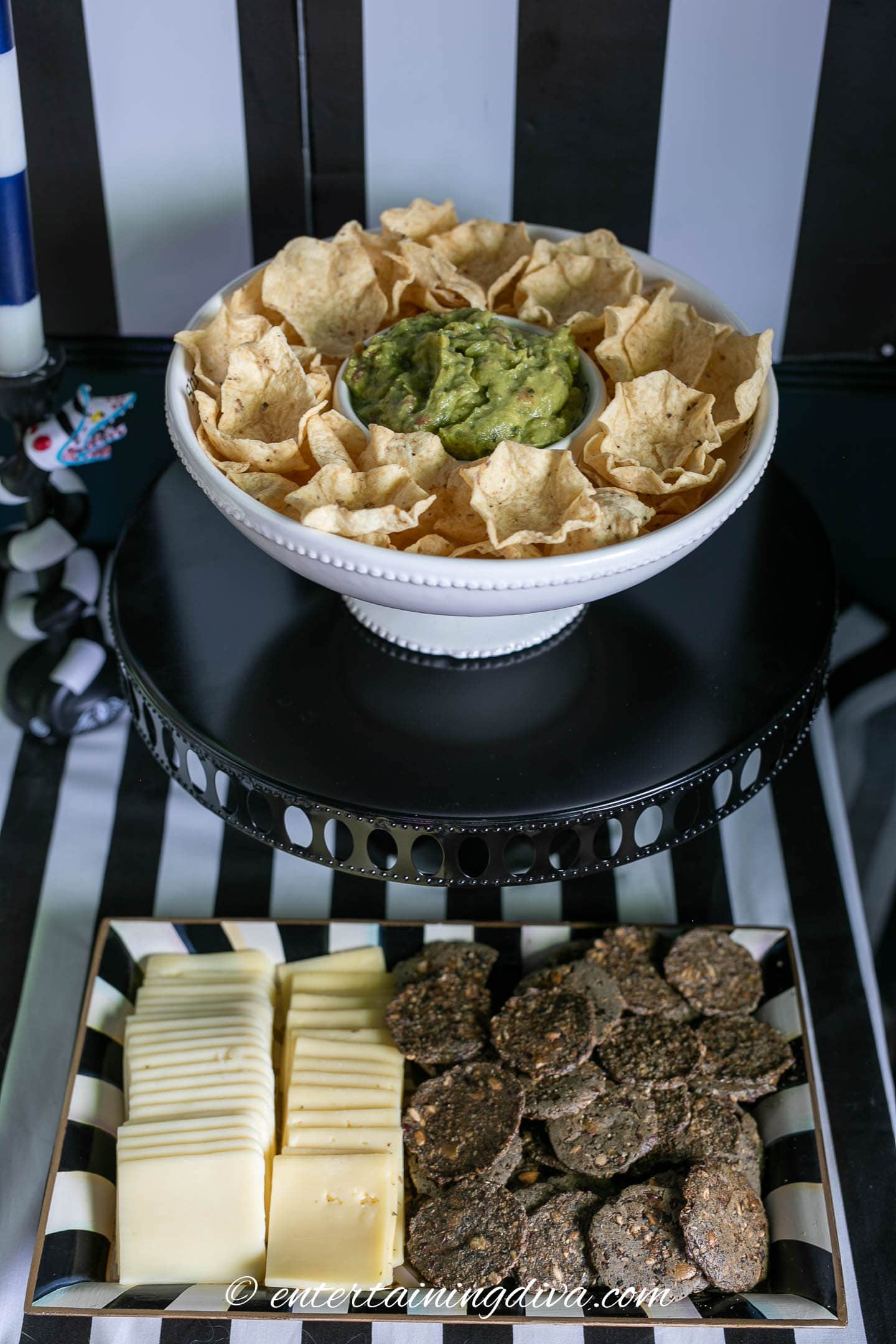Guacamole and chips in a bowl above a plate of white cheese slices and black crackers at a Beetlejuice party.