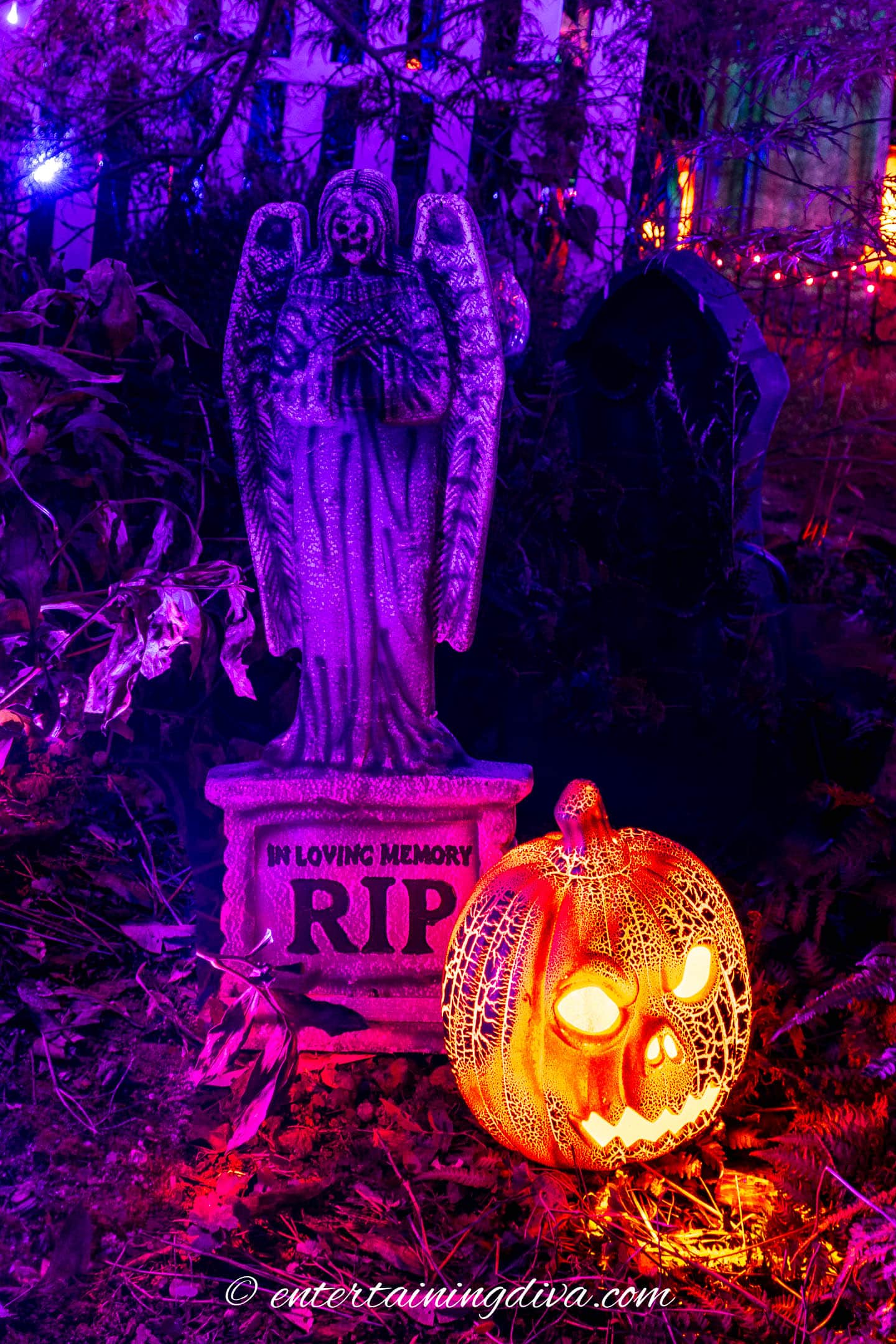 An outdoor lighted pumpkin in front of a Halloween tombstone at night.