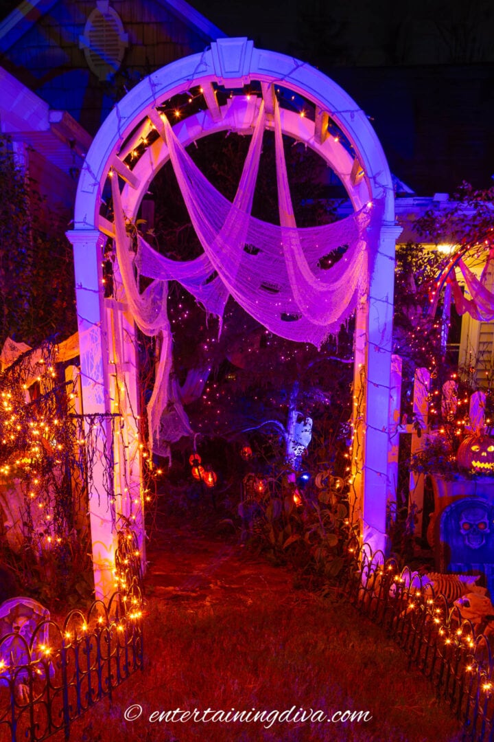 Front yard arbor decorated with white creepy cloth and lit with outdoor Halloween flood lights in blue, purple and pink