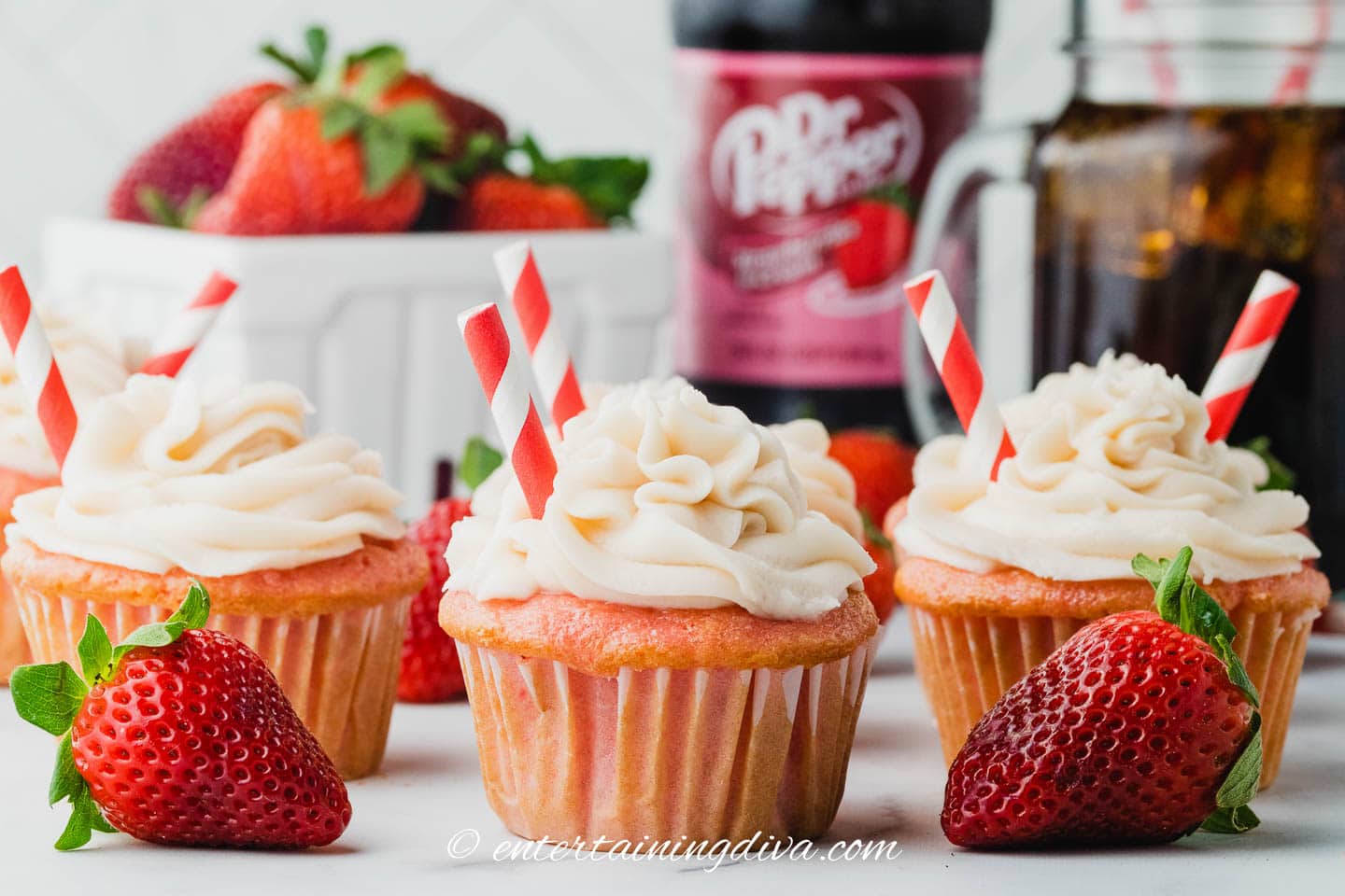 strawberry soda cupcakes in front of a basket of fresh strawberries, and a bottle of Dr Pepper