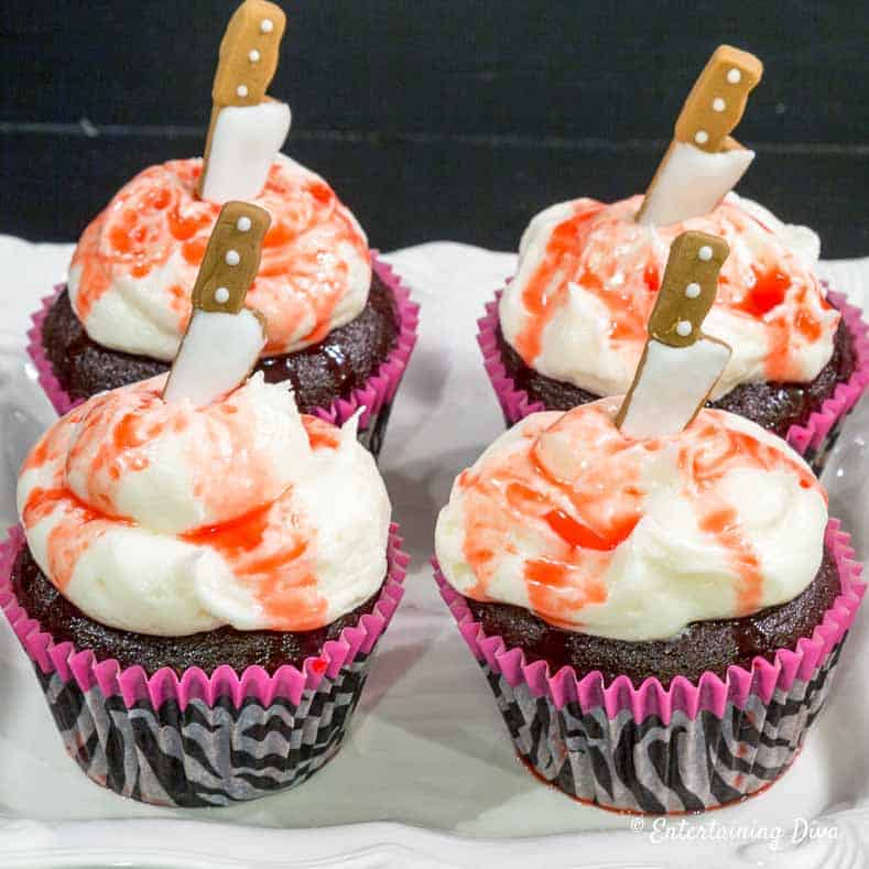 Chocolate cupcakes with white and red swirled frosting, each topped with a candy knife decoration, displayed in pink and black cupcake liners.