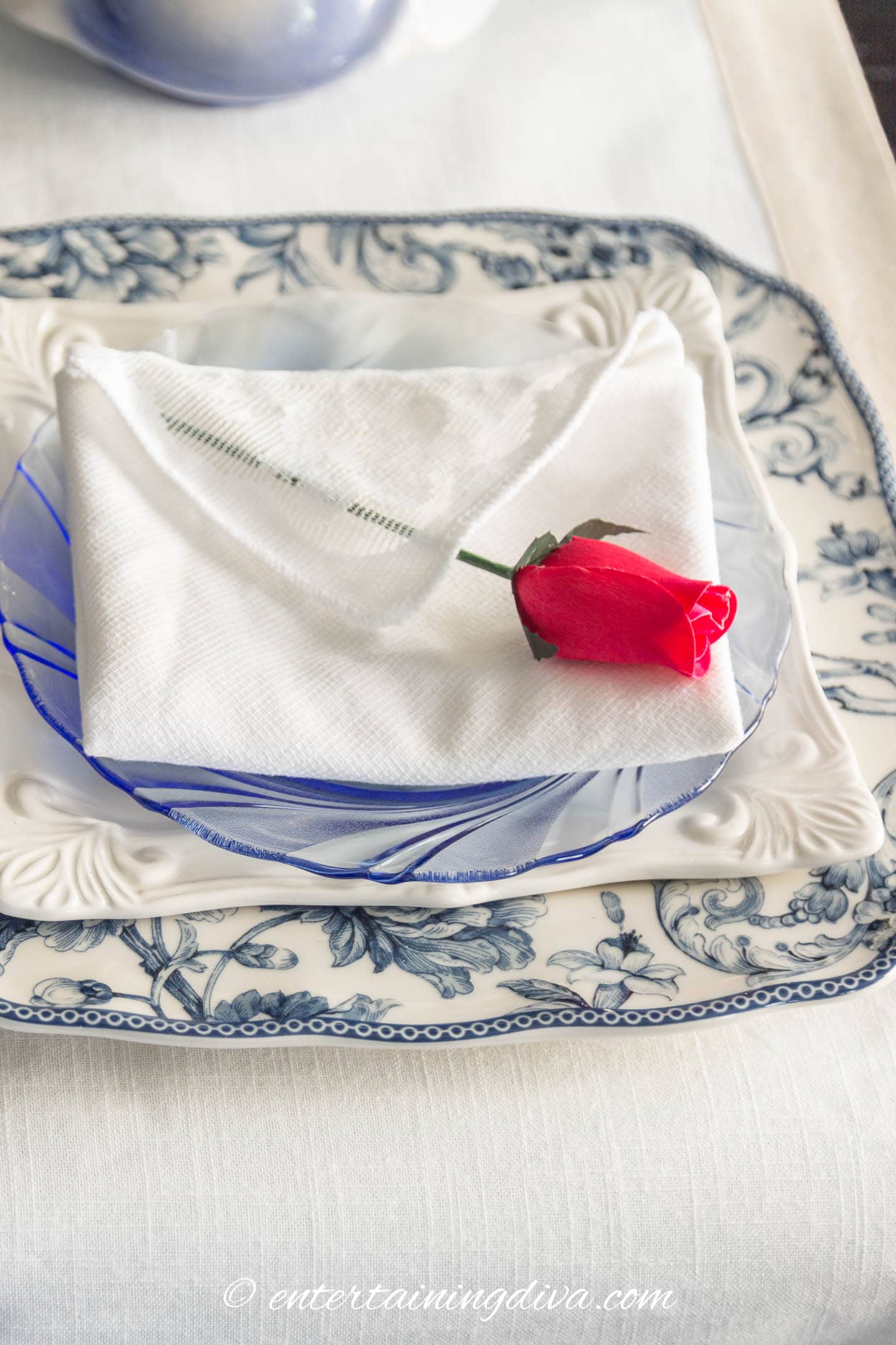 A blue and white table setting with a napkin folded into an envelope topped with a red rose
