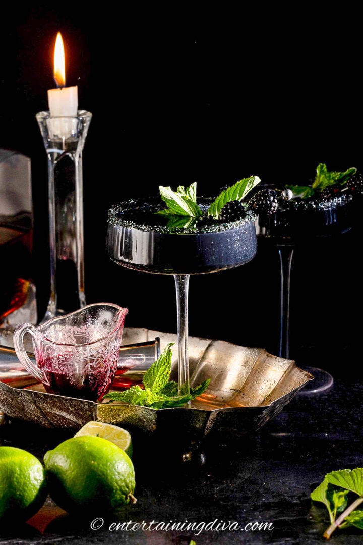 Two black cocktails garnished with mint leaves sit on a tray with a glass pitcher of dark liquid, a cut lime, and a lit candle in the background.