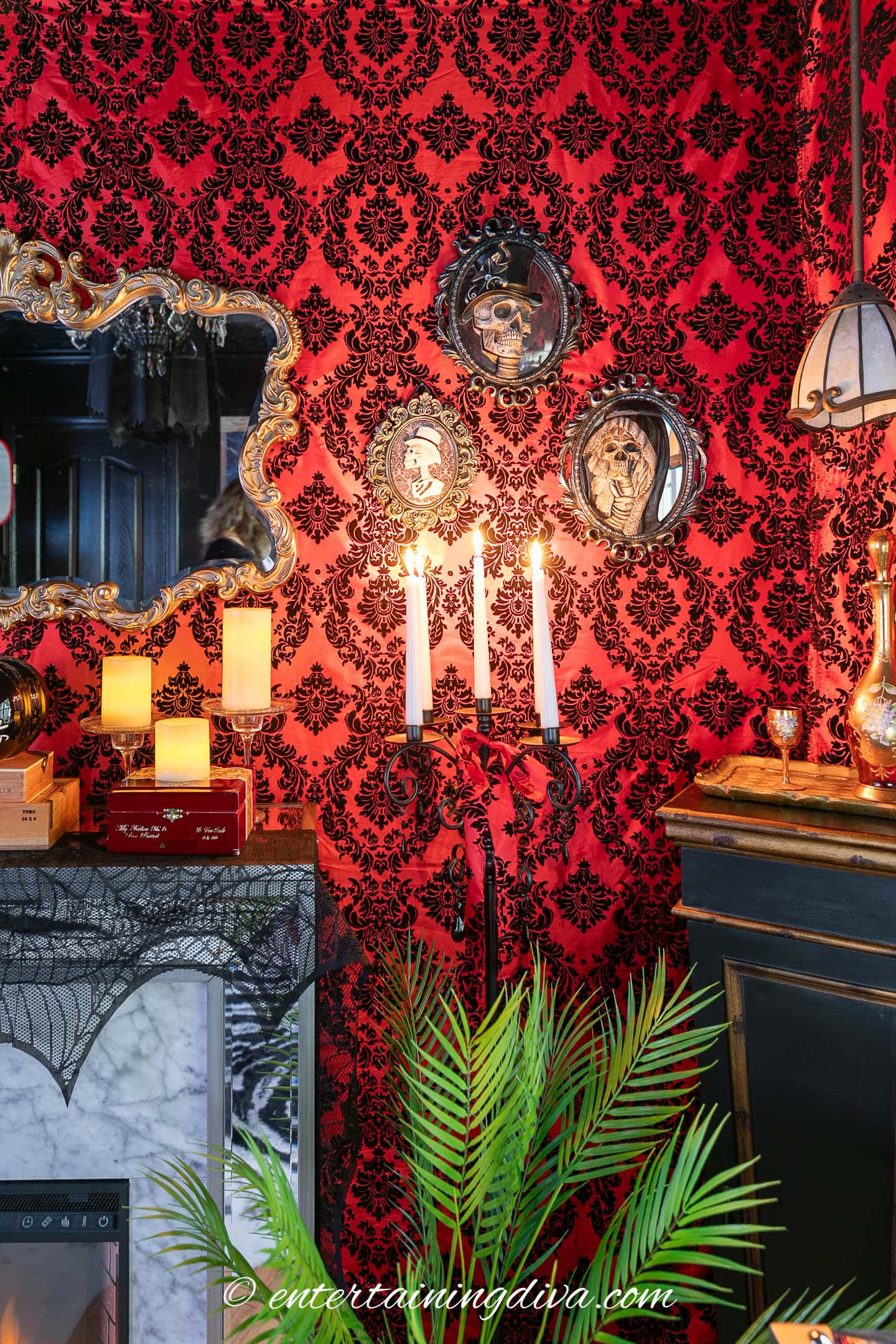 A room decorated with red damask wallpaper, ornate mirrors, lit candles, gothic decor, and a green plant in the foreground.