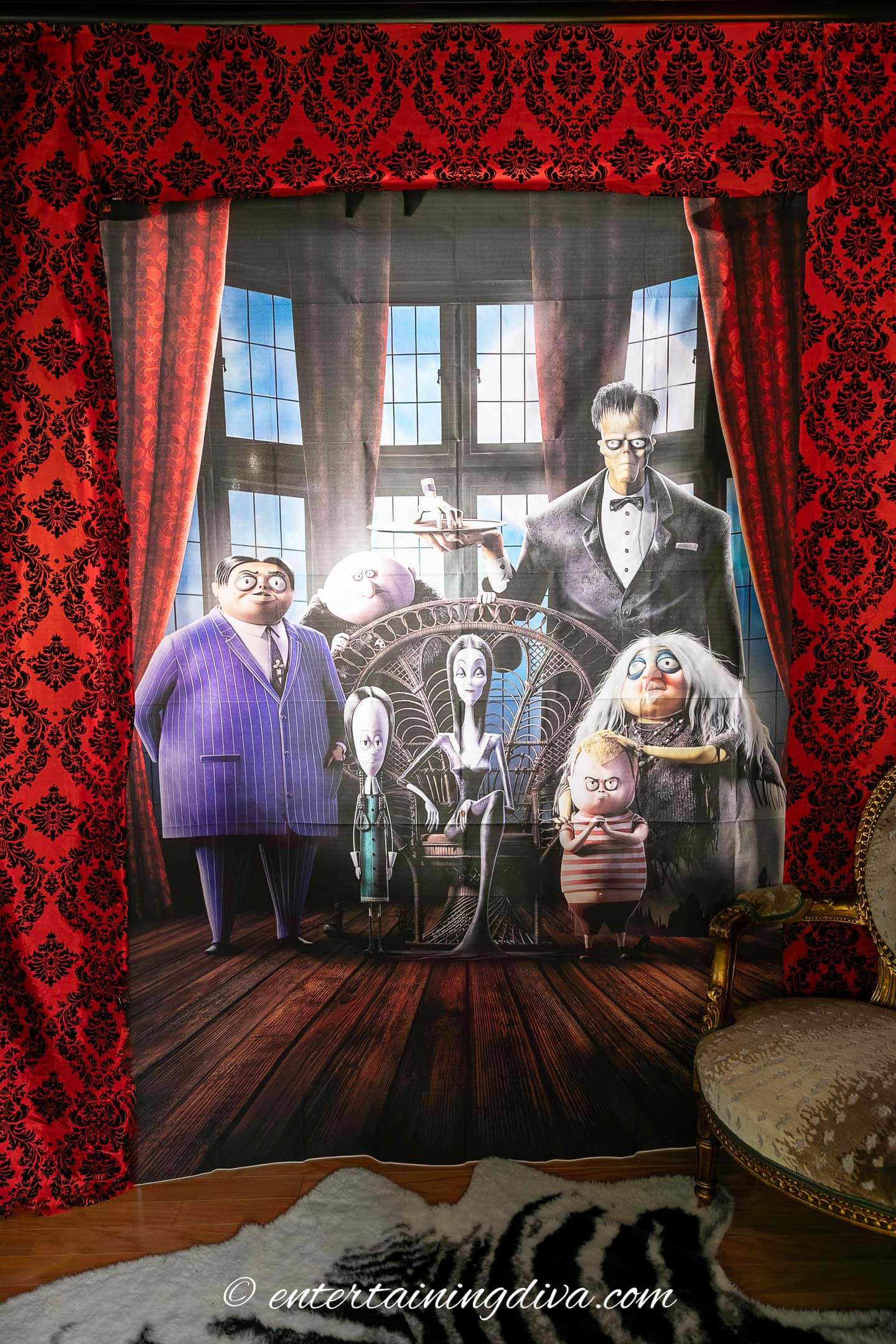 A tapestry featuring animated characters from The Addams Family stands against red damask curtains, with an ornate chair partially visible on the right.