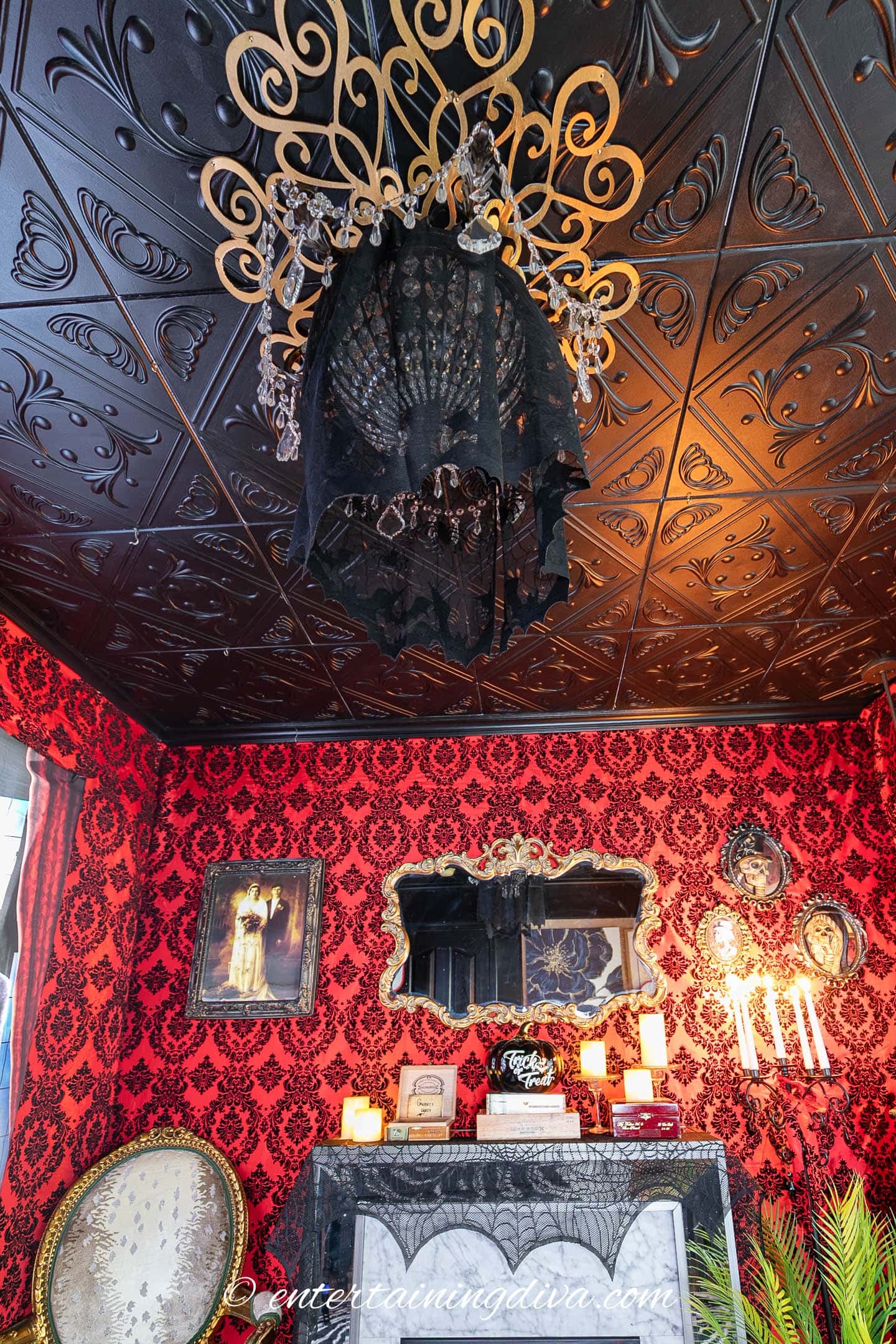 Room with ornate black tin ceiling, chandelier draped in black lace, red damask wallpaper, vintage furniture, candles, and gothic decor elements including framed art and mirror.