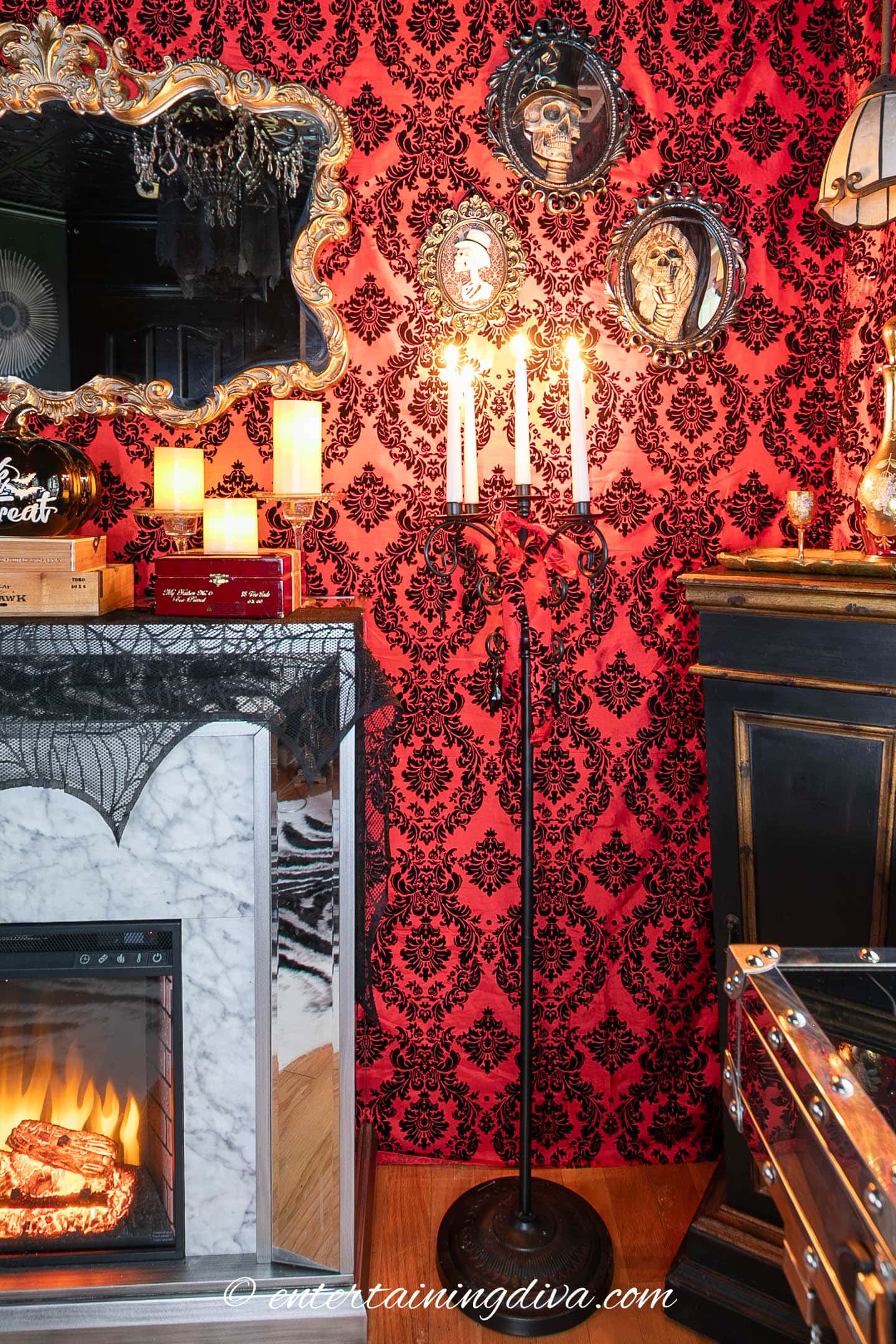 A room decorated for Halloween with a red damask wallpaper, a lit candelabra, candles, gothic mirror, skull art, and a faux fireplace with spider web accents.