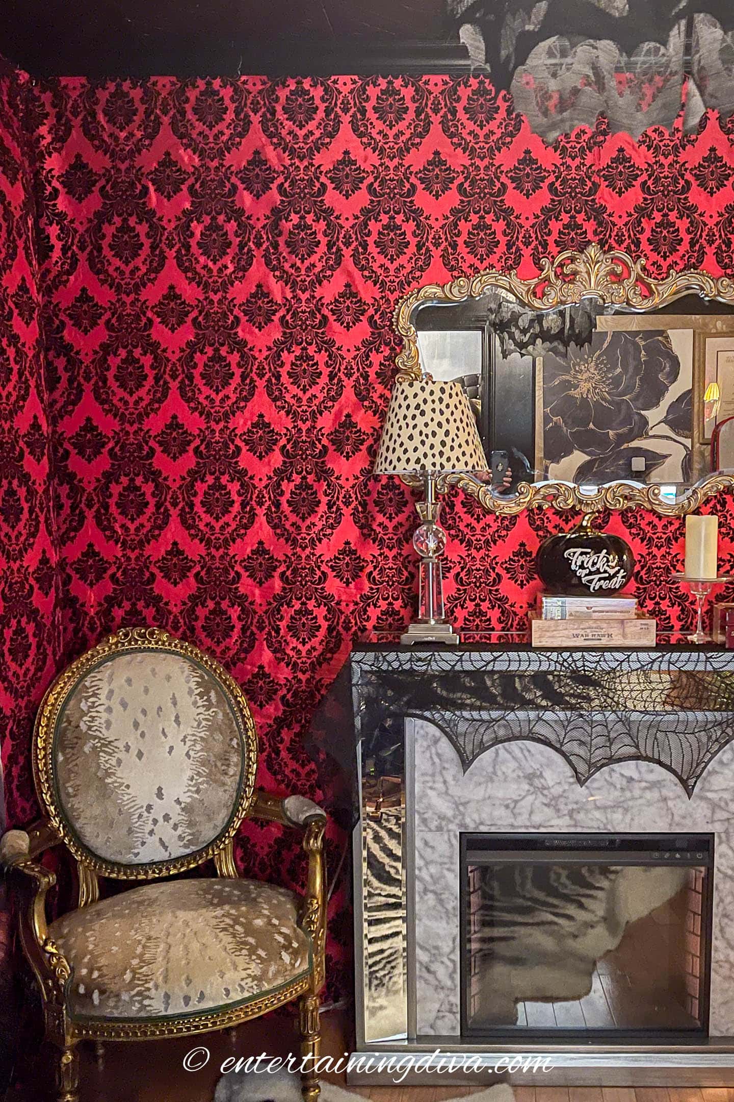 A room with ornate red and black wallpaper, an antique armchair, a marble fireplace, a decorative mirror, and Halloween-themed accents like faux cobwebs.