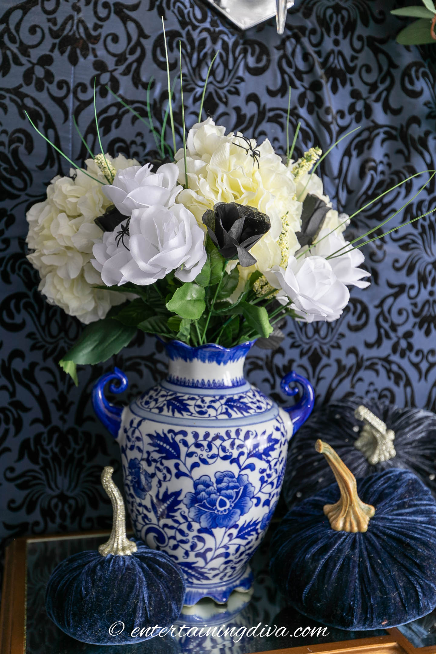 A blue and white floral vase holds white and black artificial flowers, flanked by two blue velvet pumpkins with gold stems, against a dark patterned background.