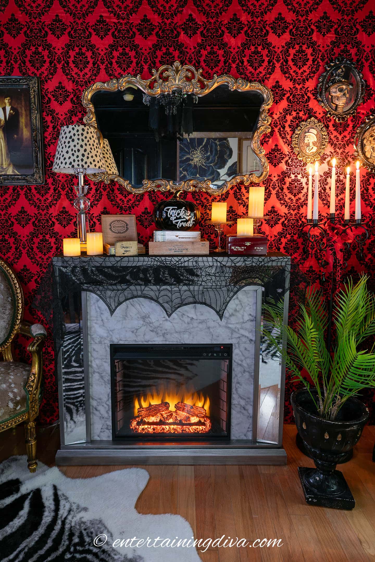 A fireplace with faux flames is decorated with cobwebs, candles, books, and a skull, set against a red damask wall with a mirror, framed art, and candelabra.