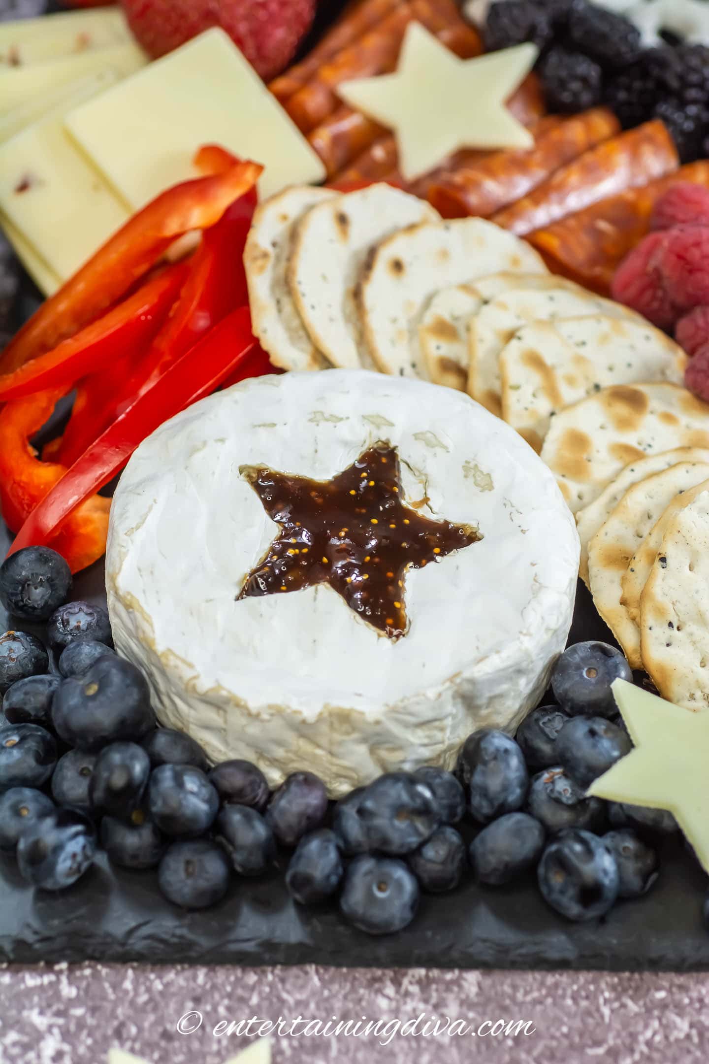 red, white and blue charcuterie board with blueberries, white cheese, crackers, red peppers and raspberries