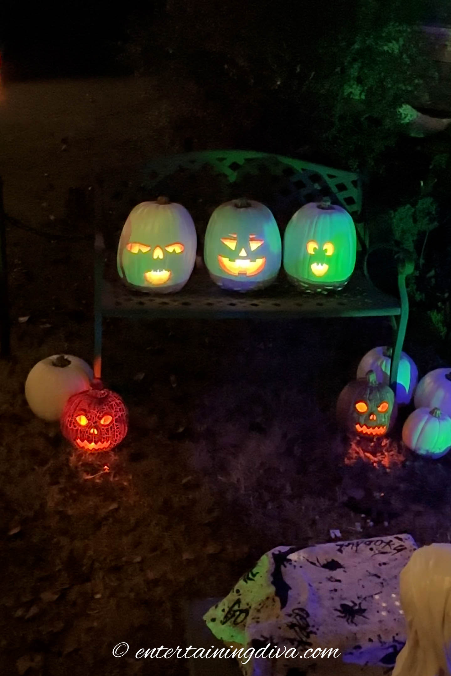 A group of glowing carved pumpkins with various facial expressions are displayed on a bench and ground, surrounded by white pumpkins at night.