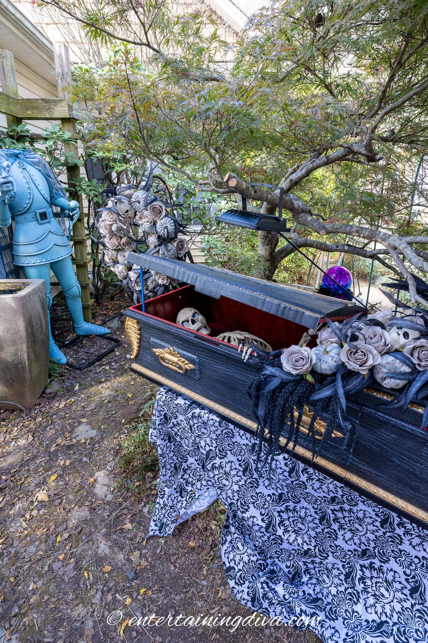 A skeleton lies in an open black coffin adorned with gray flowers in a garden, next to a blue suit of armor and patterned fabric on the ground.