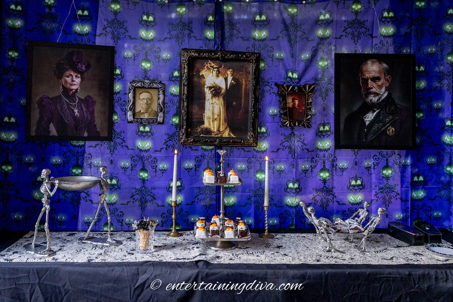 A Halloween-themed table is set with skeleton decorations, potion bottles, and candles in front of a blue wall with spooky portraits and glowing eyeball patterns.