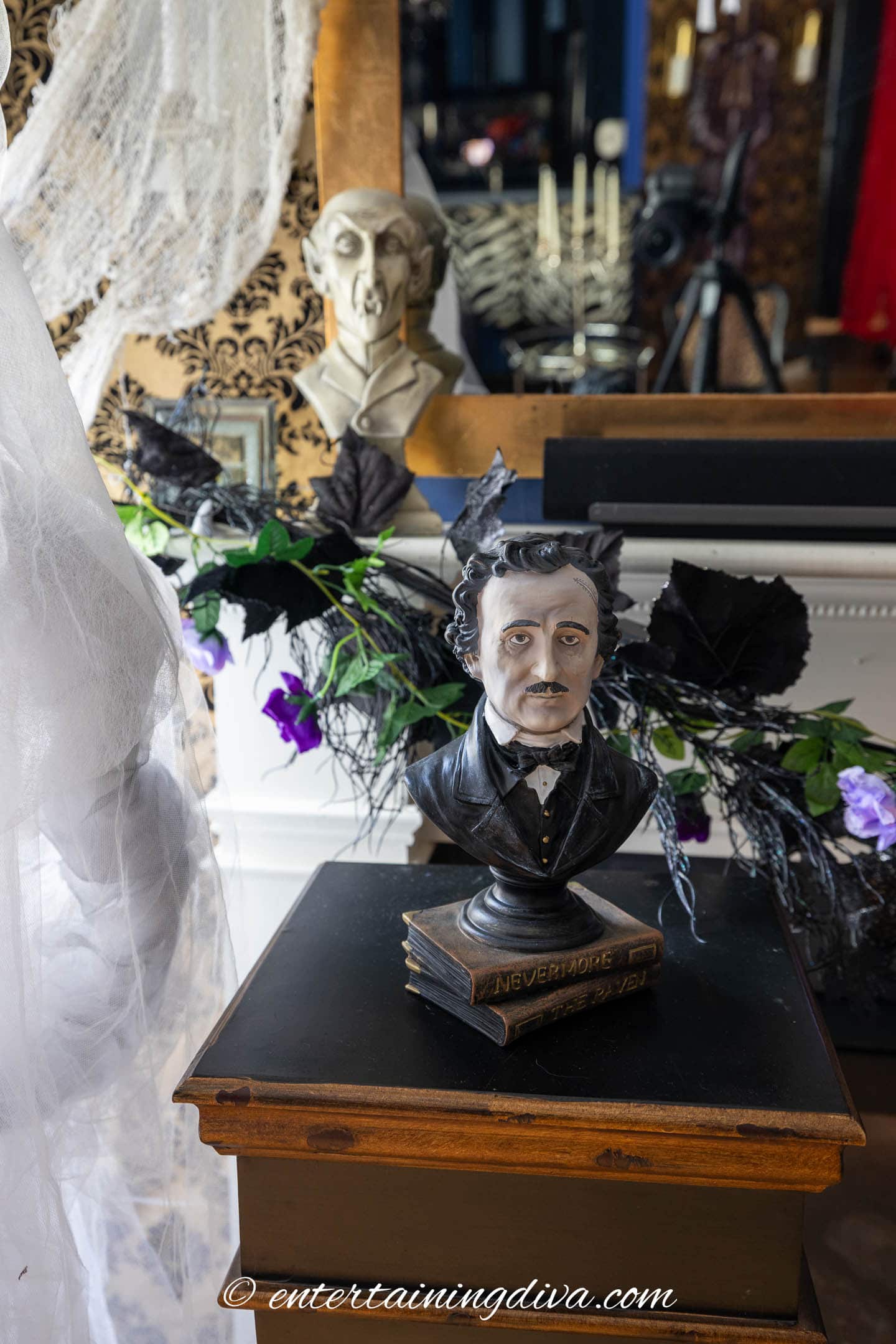 A bust of Edgar Allan Poe labeled “Nevermore,” sits on a small table in front of dark foliage and flowers, with a draped white fabric and another bust in the background.