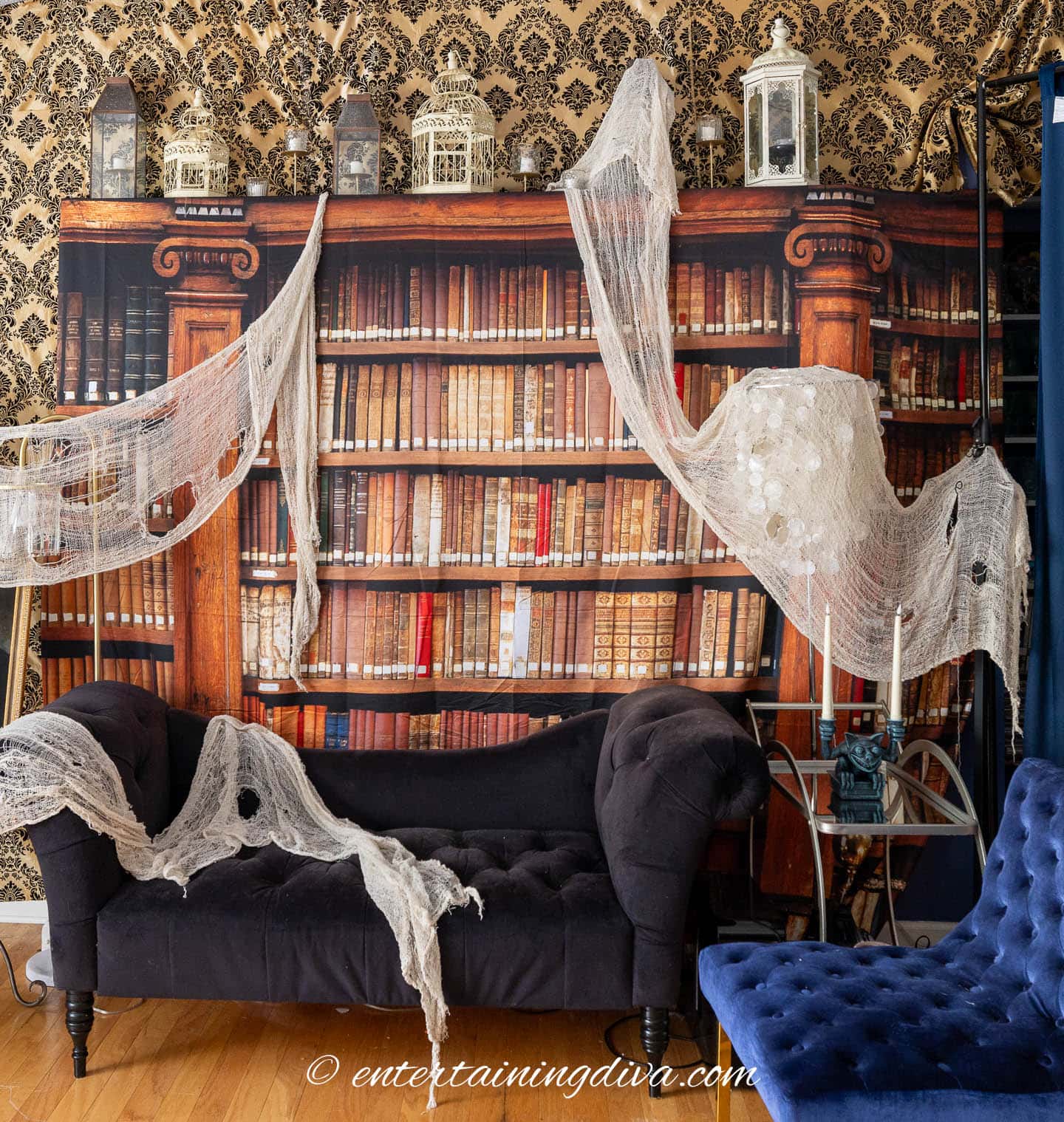 A room with a black couch, faux bookshelf wallpaper, vintage lanterns, and white tattered fabric draped as Halloween decor.