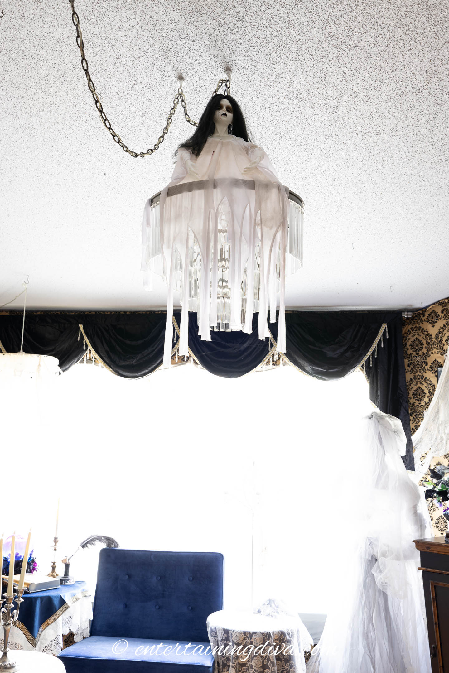 A chandelier is decorated with a ghostly figure draped in white cloth, resembling a Halloween decoration, hanging from the ceiling in a living room.