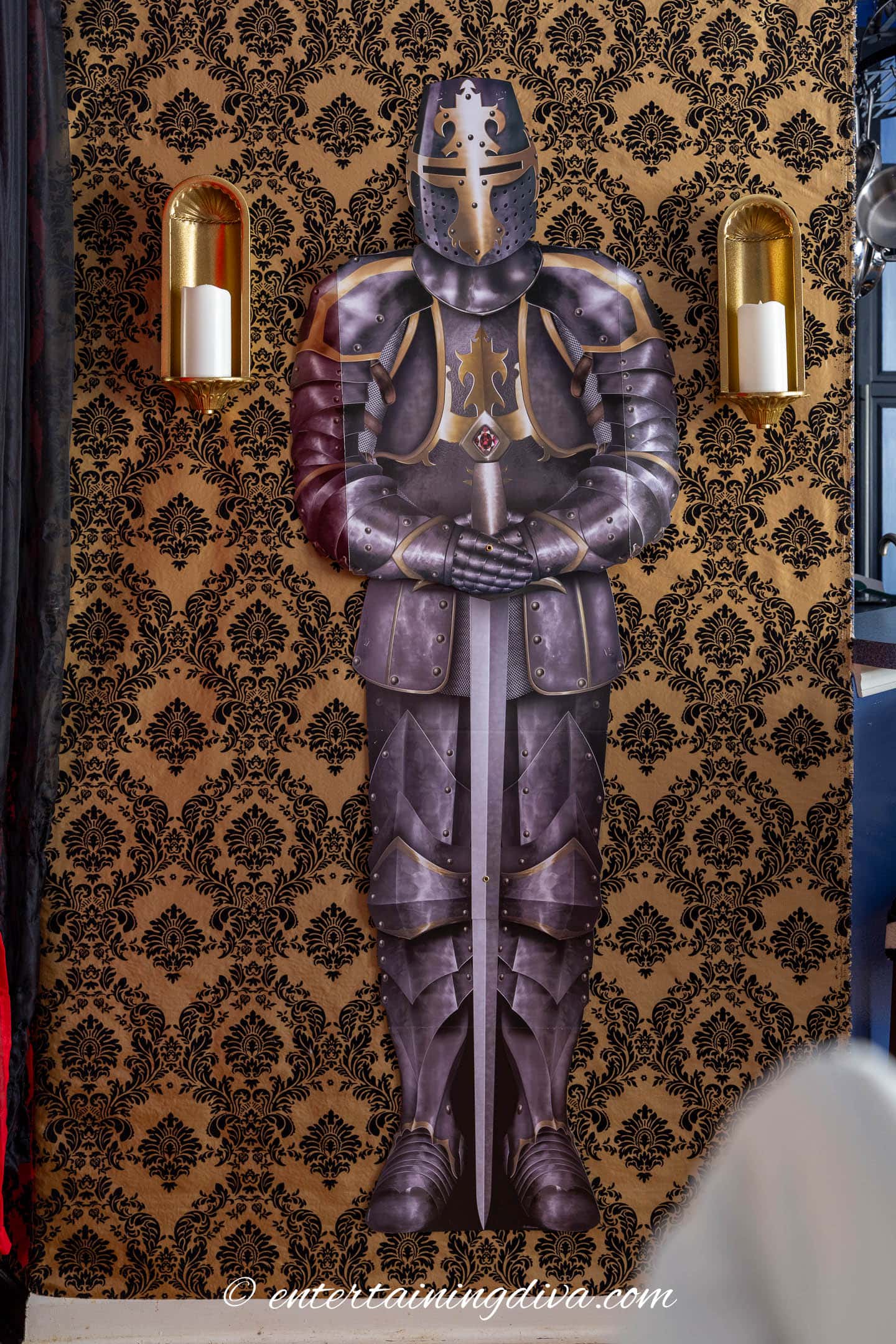 A life-size knight in armor decoration is displayed on a gold and black patterned wall, flanked by two wall sconces holding white candles.