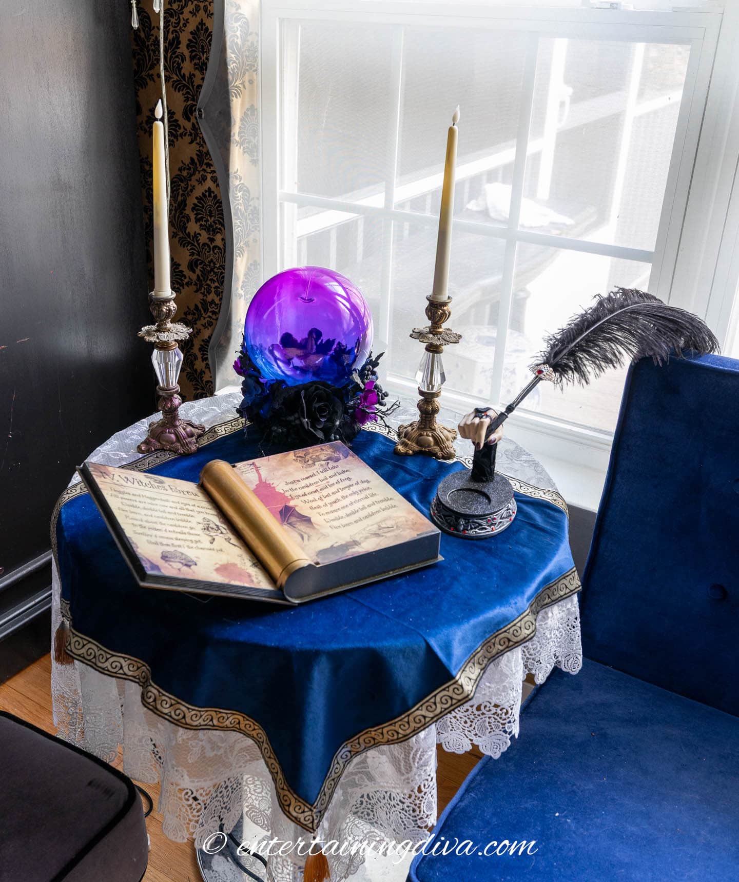 A round table with a blue cloth holds an open book, a gold scroll, a purple crystal ball, two candlesticks, and a quill in an inkwell near a window.