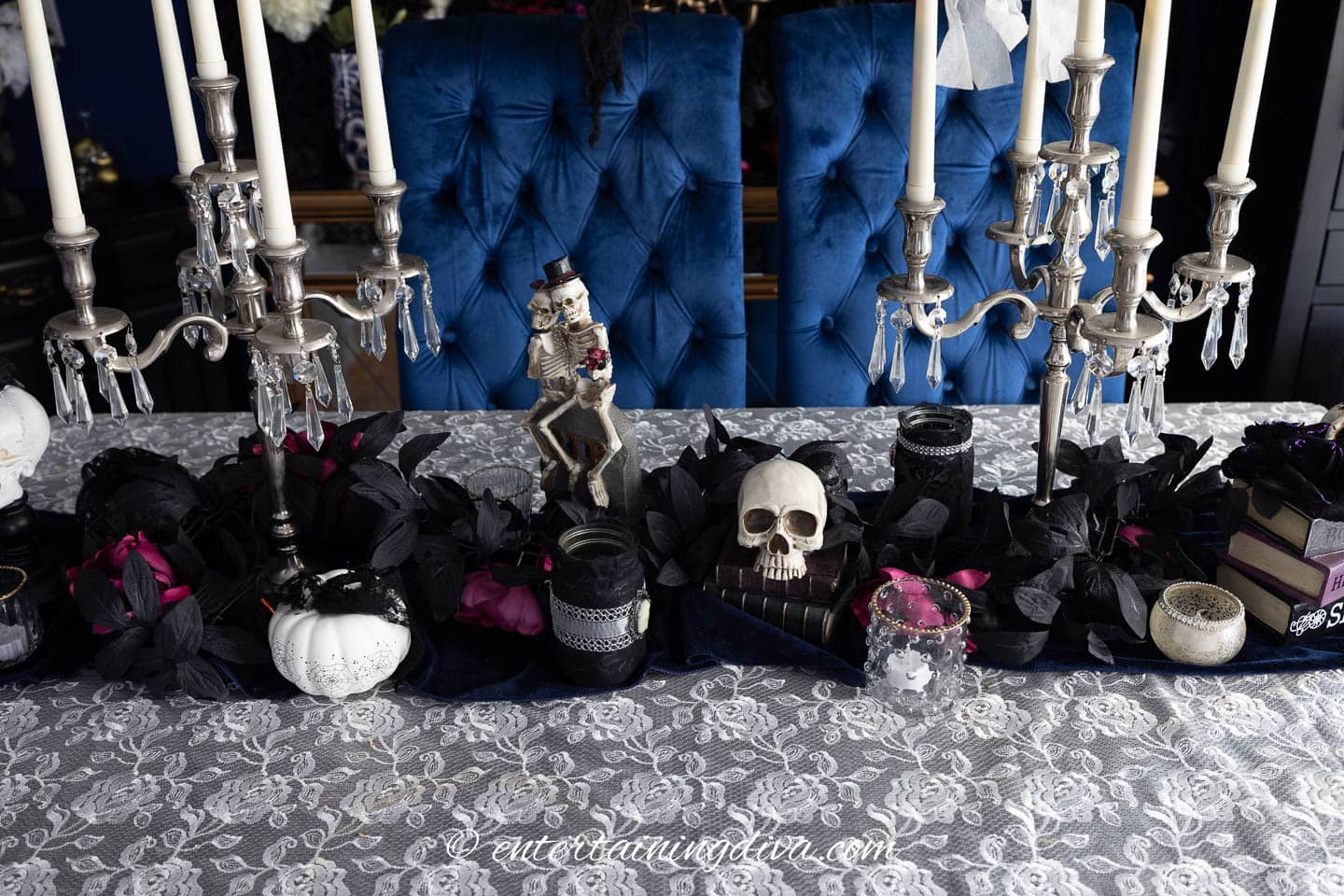 A dining table with a lace tablecloth is decorated with black leaves, candles, a white pumpkin, small skulls, and a skeleton couple figurine.