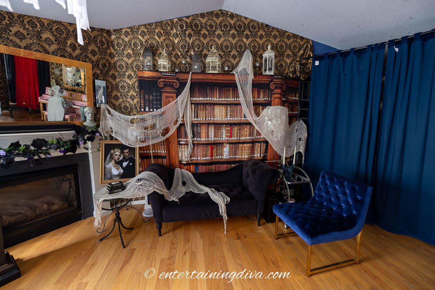 A sitting room decorated with Halloween gauze, vintage bookshelves, a fireplace, dark curtains, and a blue velvet chair.