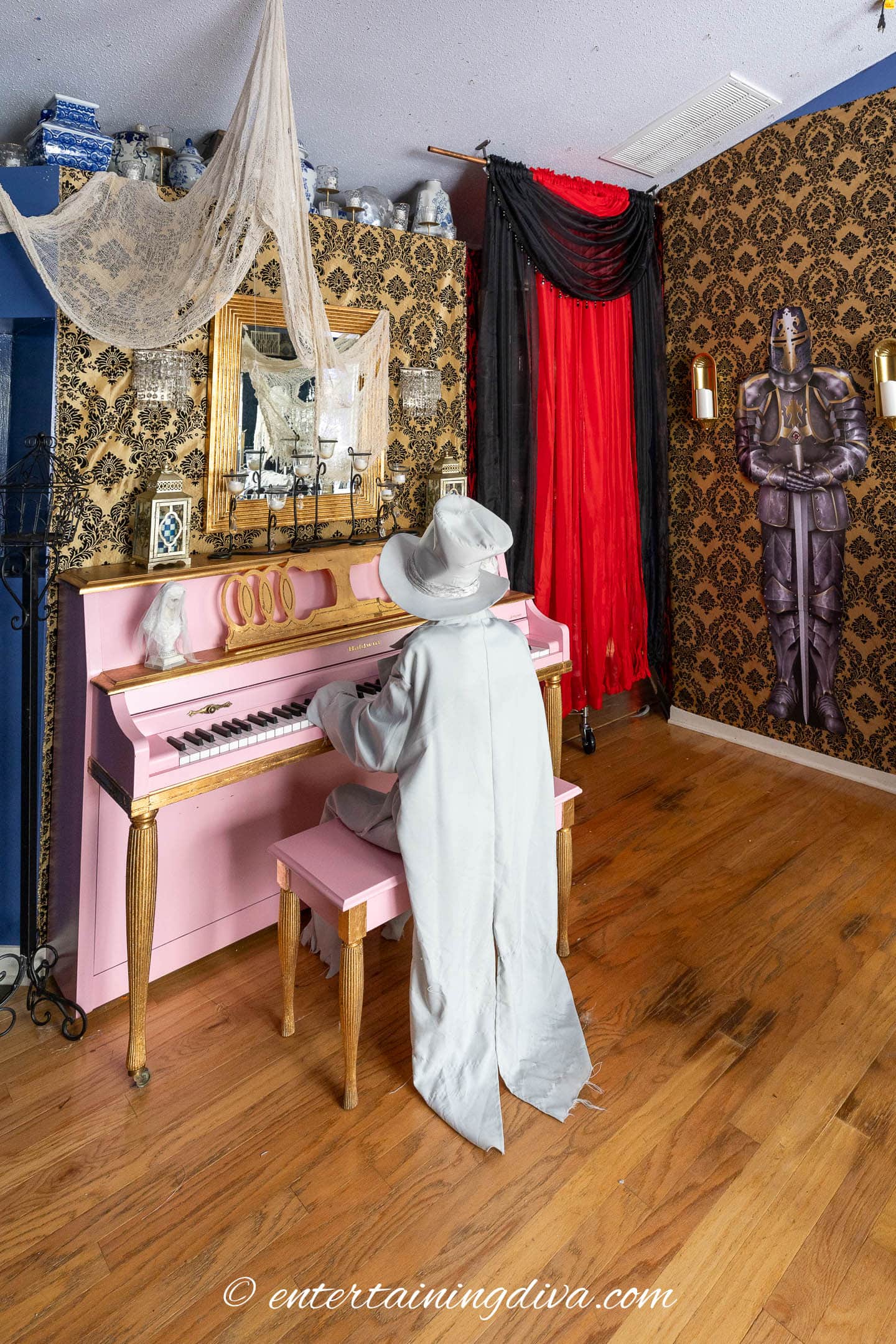 A person in a light gray wizard costume plays a pink piano in an ornately decorated room with gold wallpaper, lace, and a knight statue.