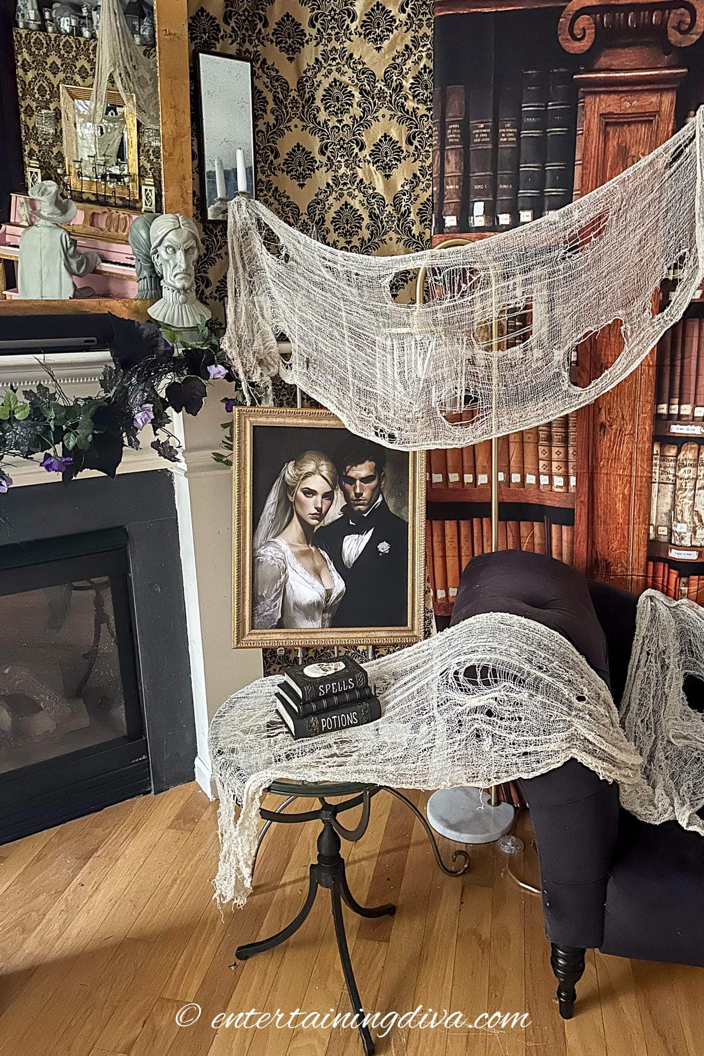 A Gothic-themed room with cobweb decorations, a fireplace, bookshelves, a dark armchair, and a framed portrait of a couple in wedding attire beside a stack of spell books.