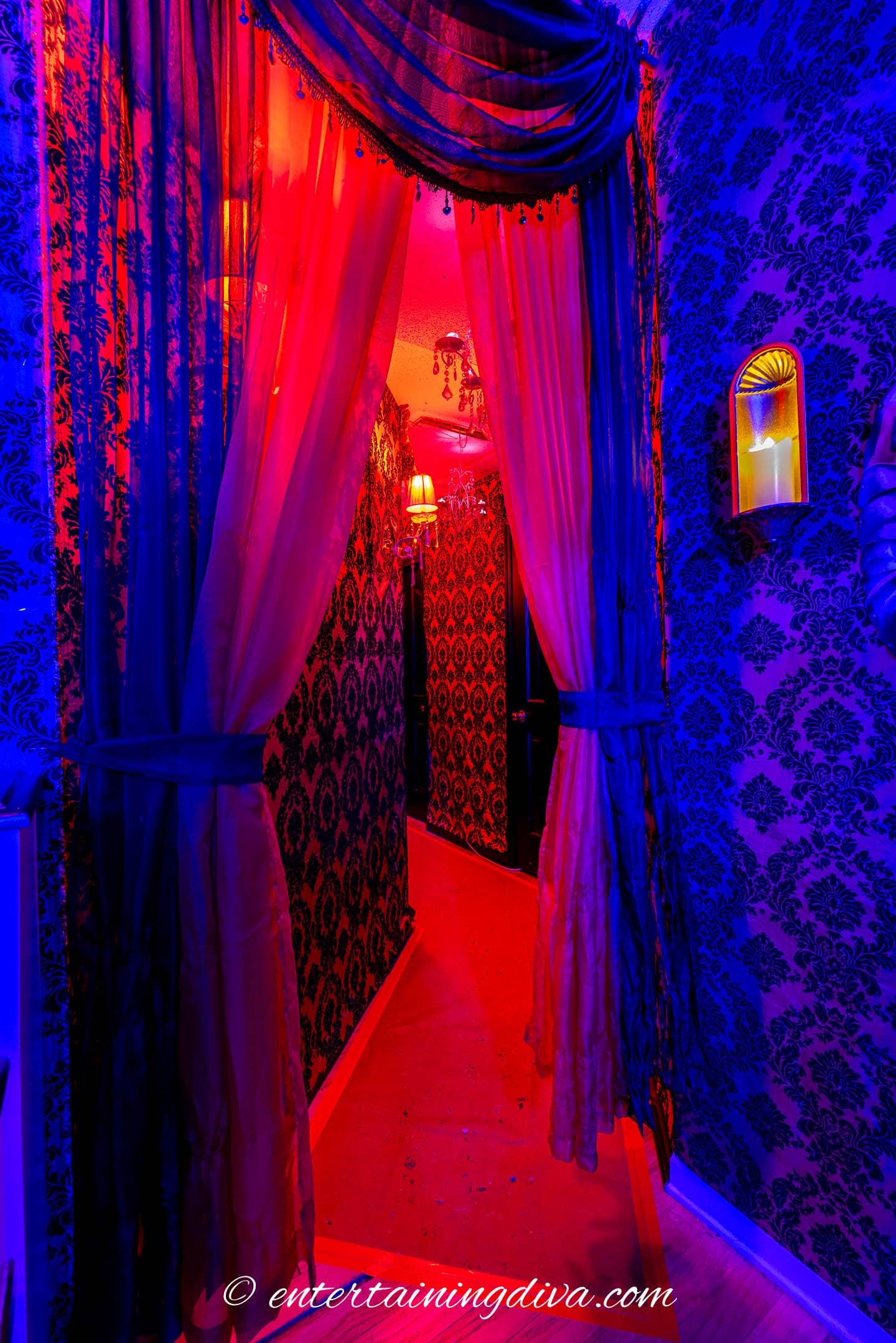 A hallway with damask wallpaper, illuminated by blue and red lights, with sheer curtains tied back and a wall sconce on the right.