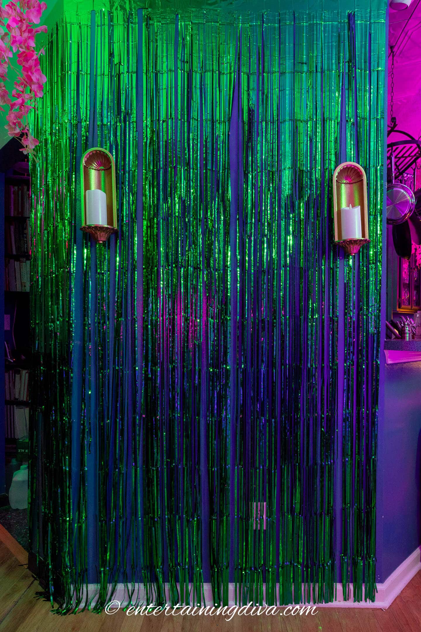 A wall decorated with green and blue metallic tinsel streamers, two candle sconces, and colorful ambient lighting.