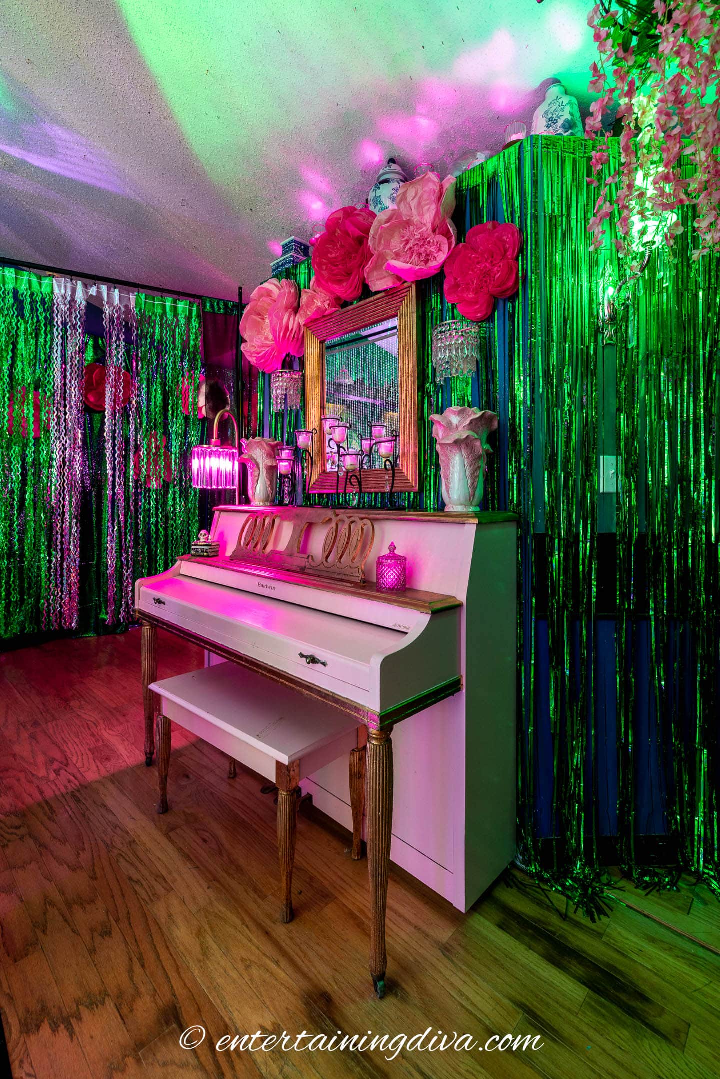 A white piano with two chairs sits against a wall decorated with green tinsel, pink flowers, and a mirror, under colorful lighting.