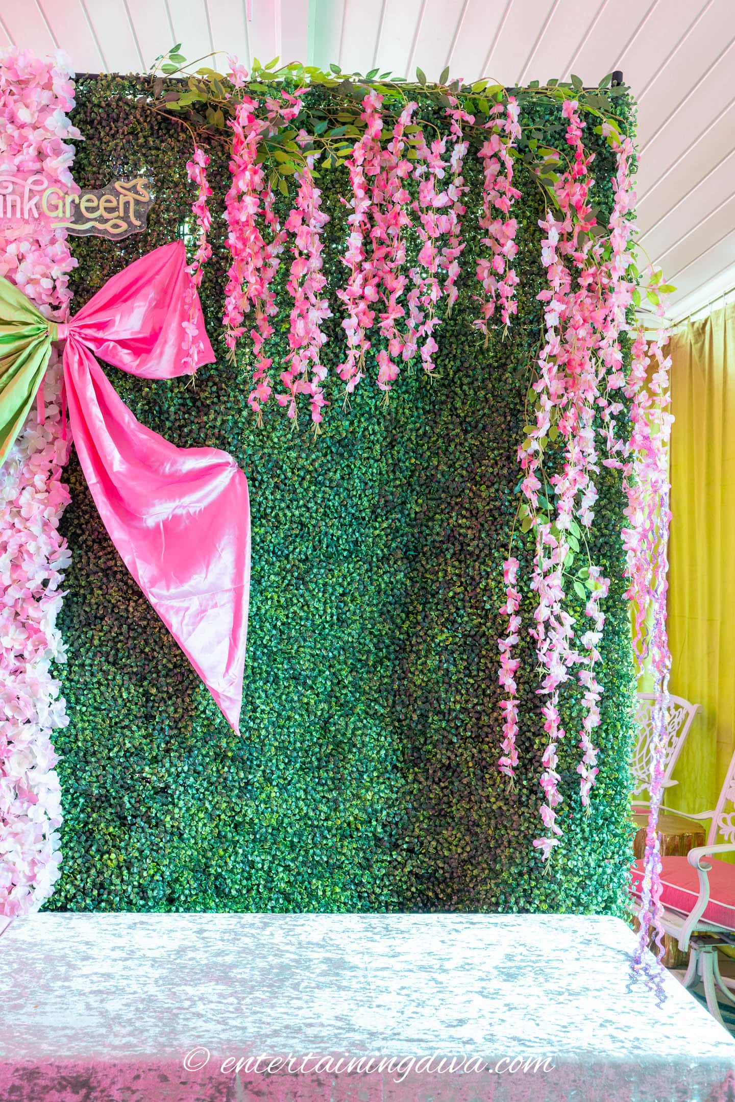 A decorative green wall with pink hanging flowers, a large pink bow, and a white floral-patterned tablecloth in front.