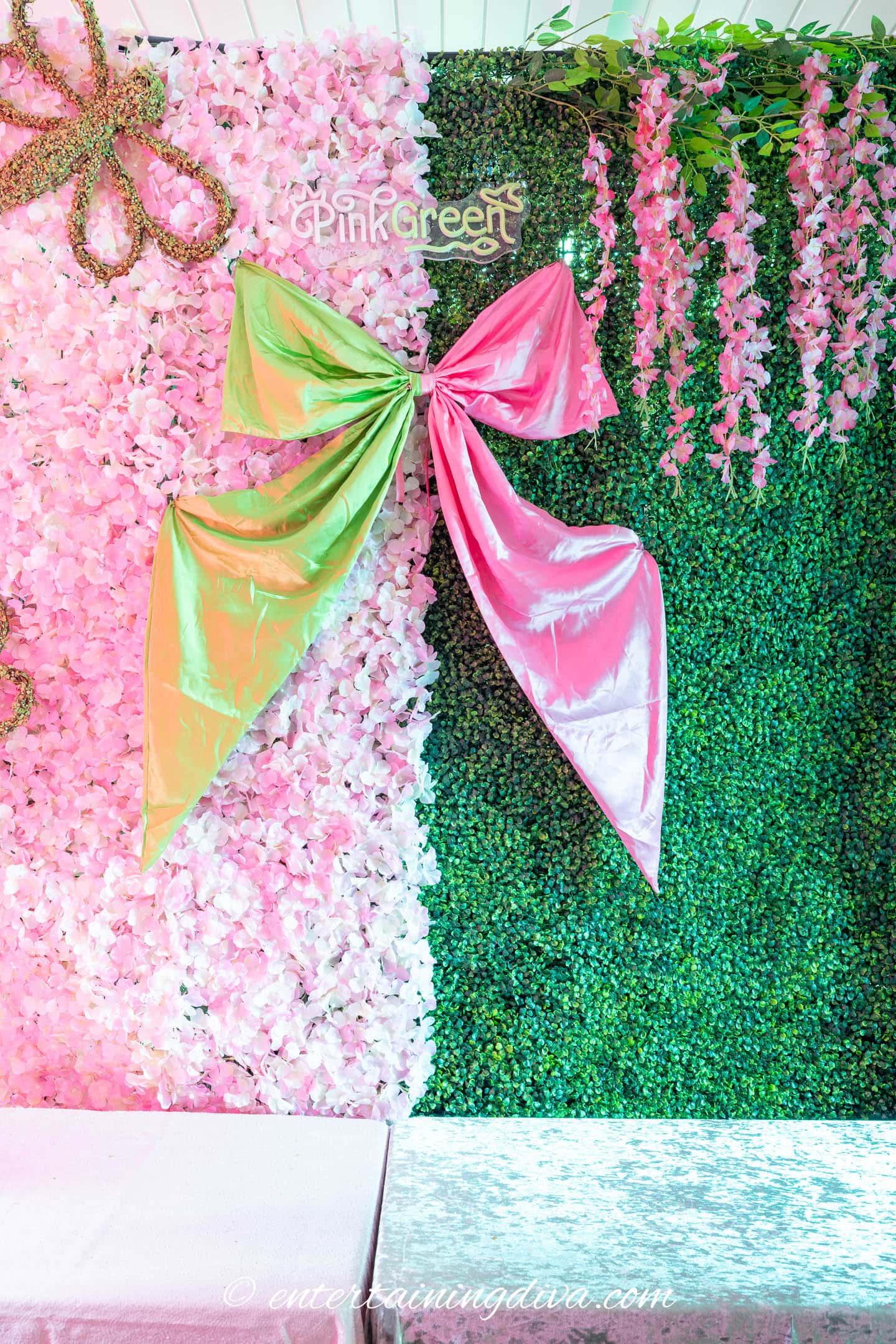 A decorative backdrop with a large metallic pink and green bow, pink artificial flowers on the left, and green foliage on the right.