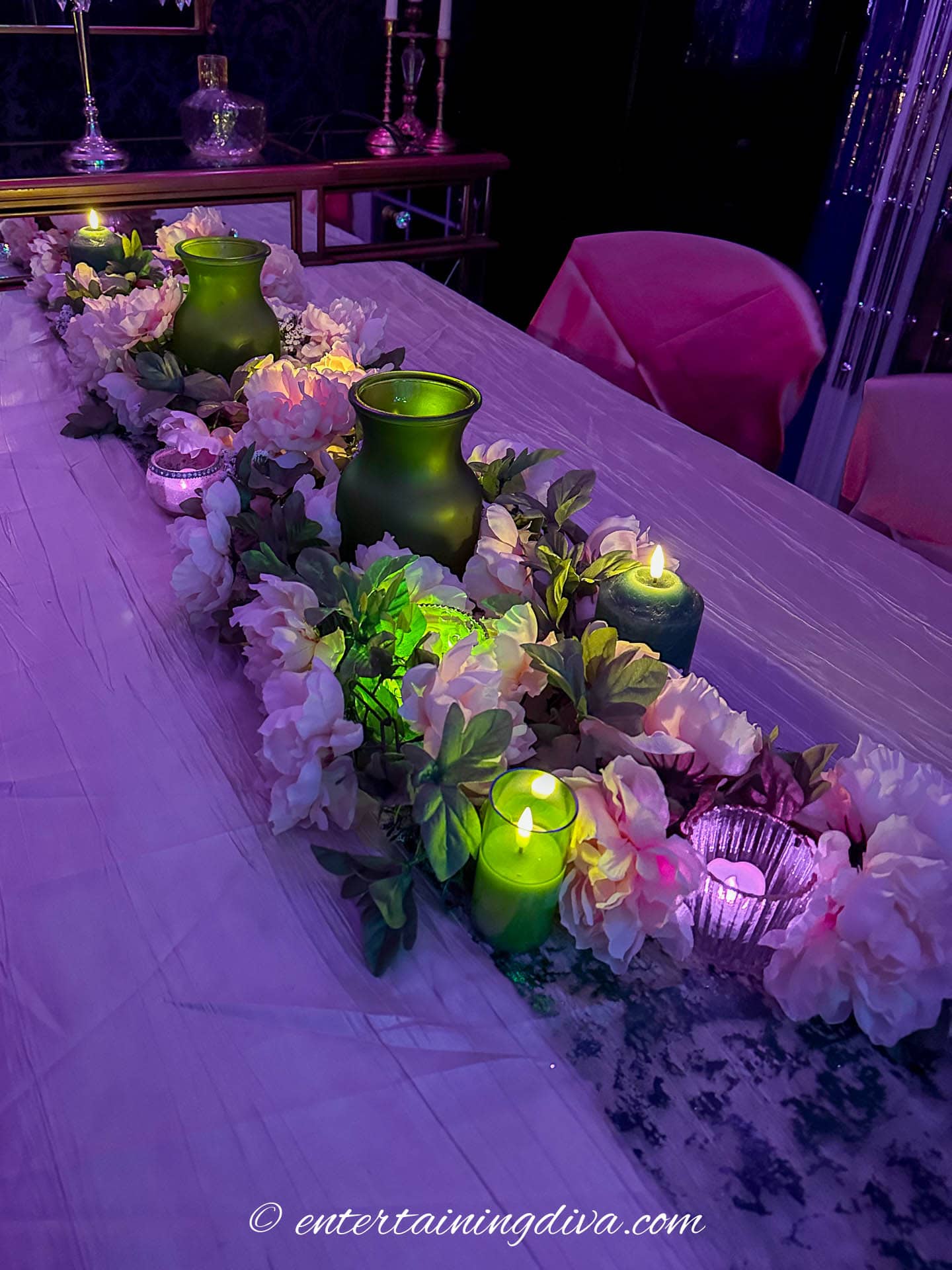 A table centerpiece with green vases, pink flowers, and lit candles is arranged on a purple-lit table.