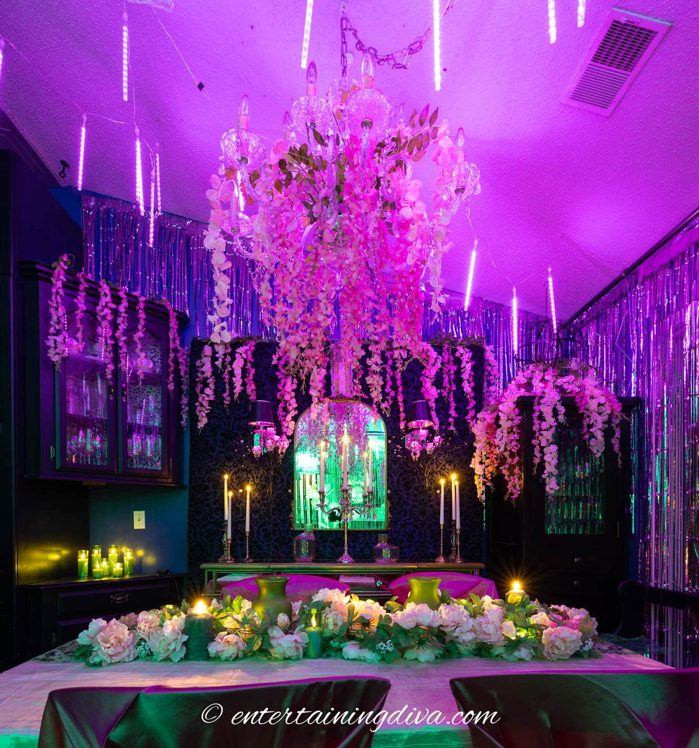 A dining room decorated for a party with purple and green lighting, floral garlands, candles, a chandelier, and hanging tinsel.