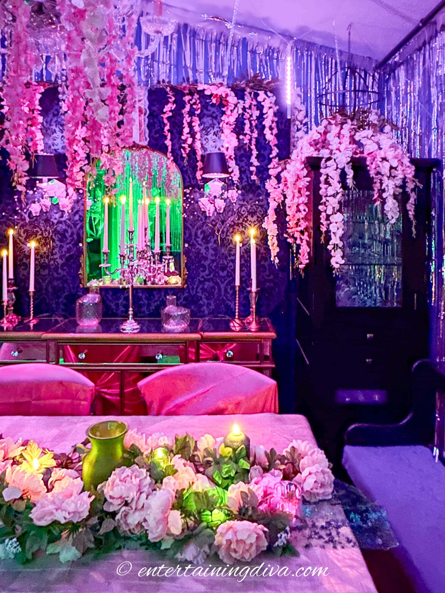 A dining room decorated with hanging pink flowers, candles, and tinsel, lit by purple and green lighting, with a floral centerpiece on the table.