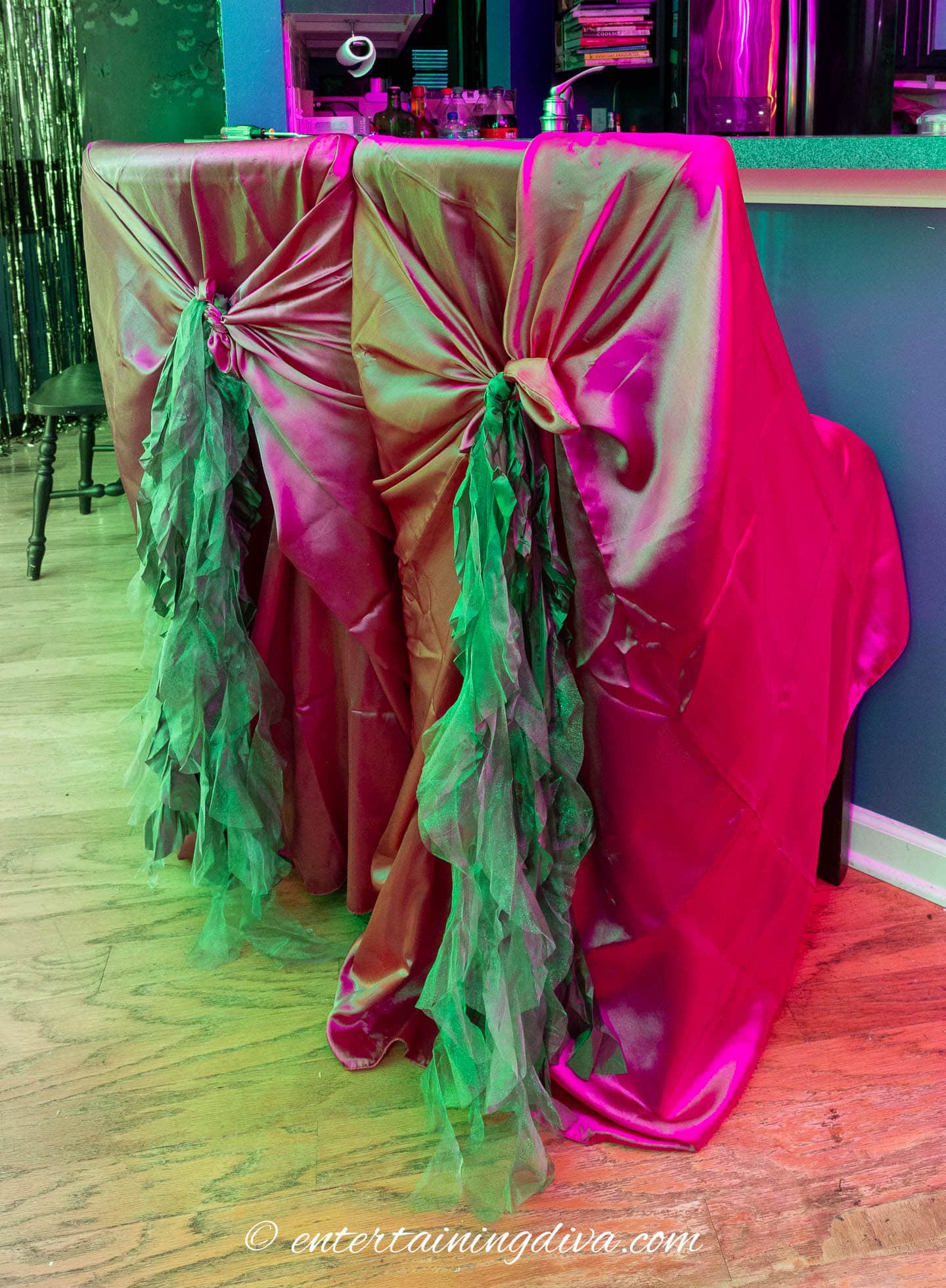 Two bar stools are covered with shiny pink fabric and decorated with green ruffled tulle bows, positioned at a counter in a colorfully lit room.