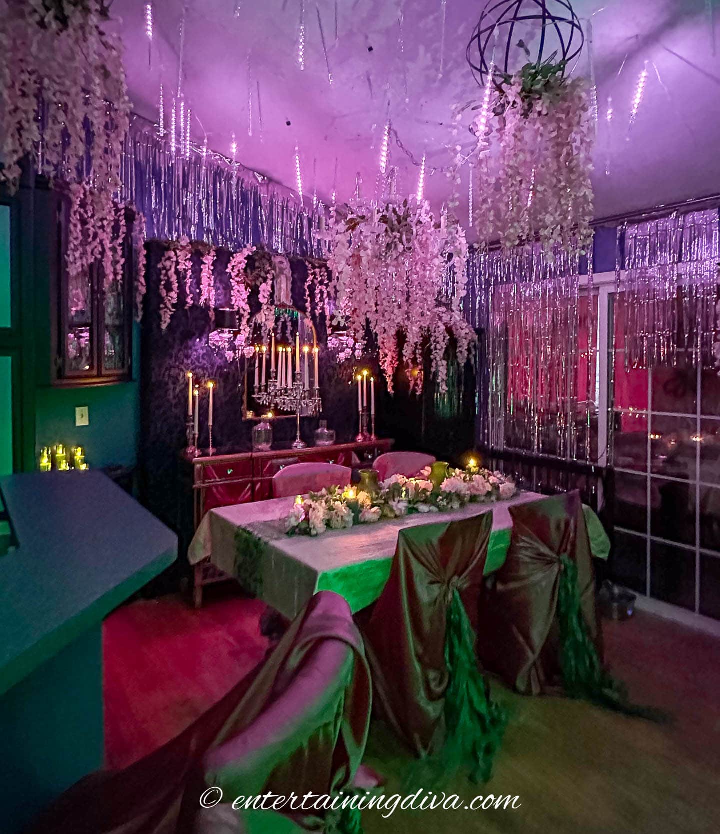 A dining room decorated with hanging flowers, draped fabric, candles, and purple lighting, creating a whimsical, enchanted forest atmosphere.